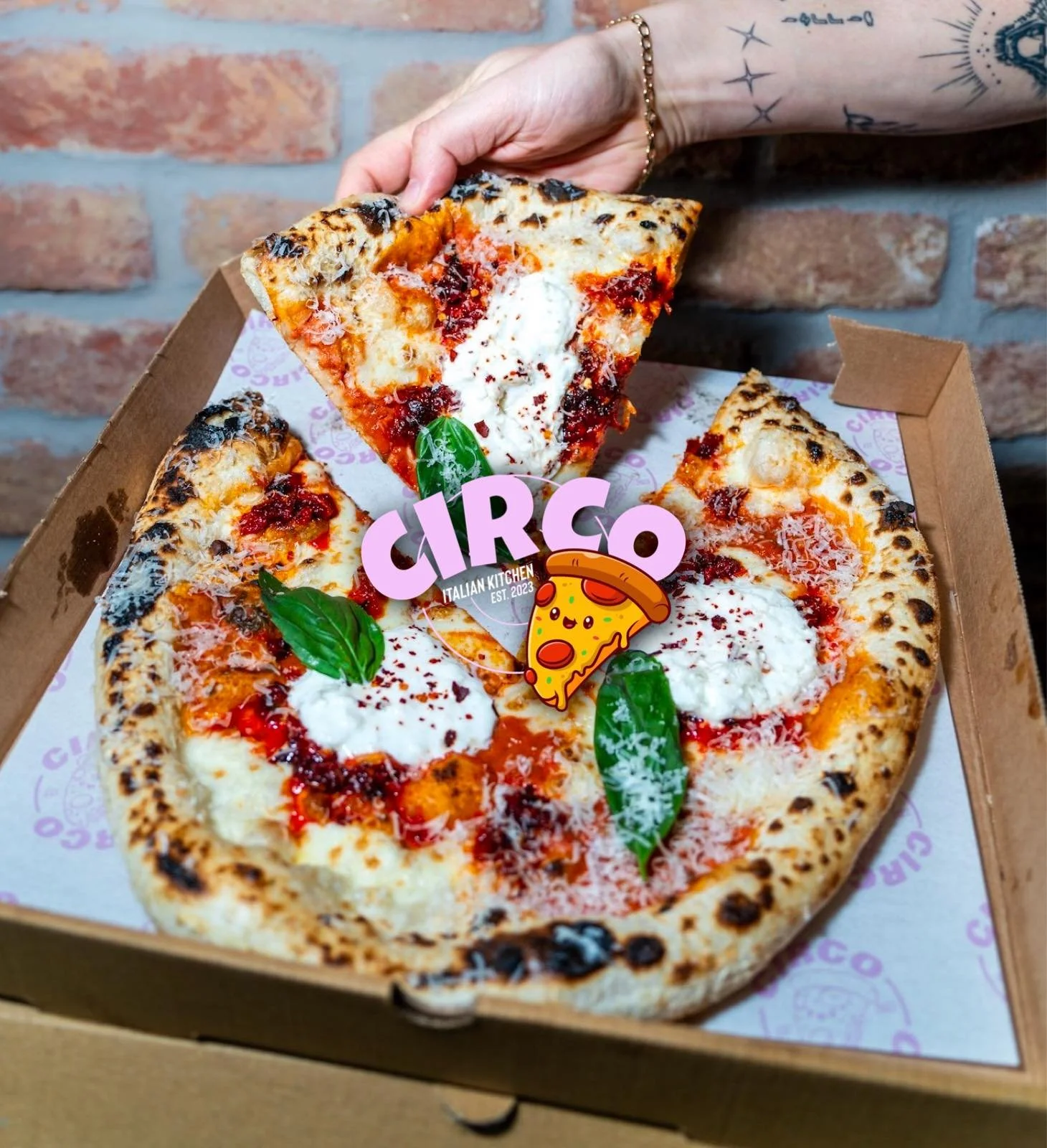 A pizza with four slices in a box, topped with tomato sauce, melted cheese, dollops of ricotta cheese, fresh basil leaves, and a sprinkle of red chili flakes. A hand is lifting one slice, and a pizza-themed sticker with the text 'CIRCO' and an illust