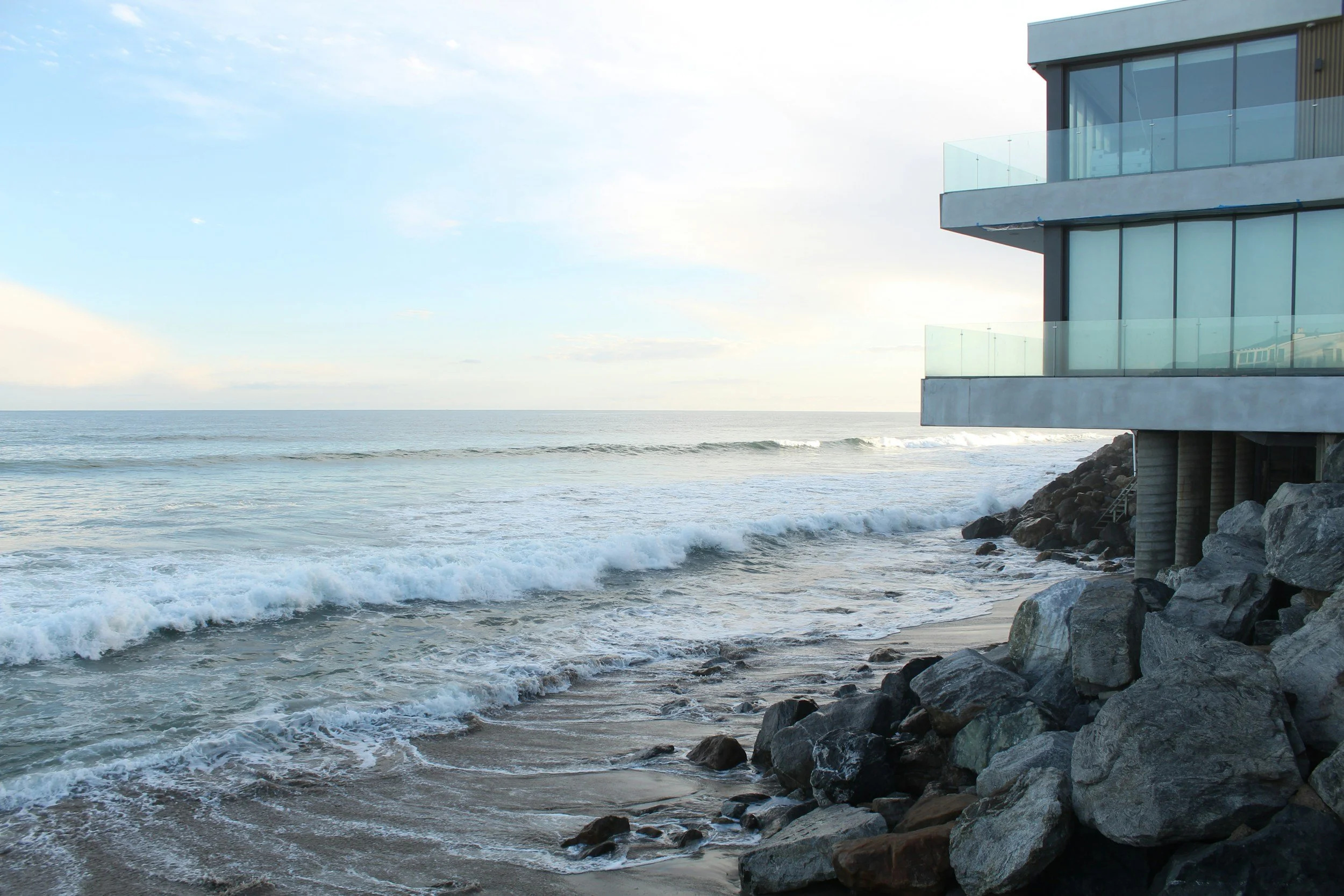 Modern building with large glass windows and balconies situated on rocky coastline with ocean waves and cloudy sky.