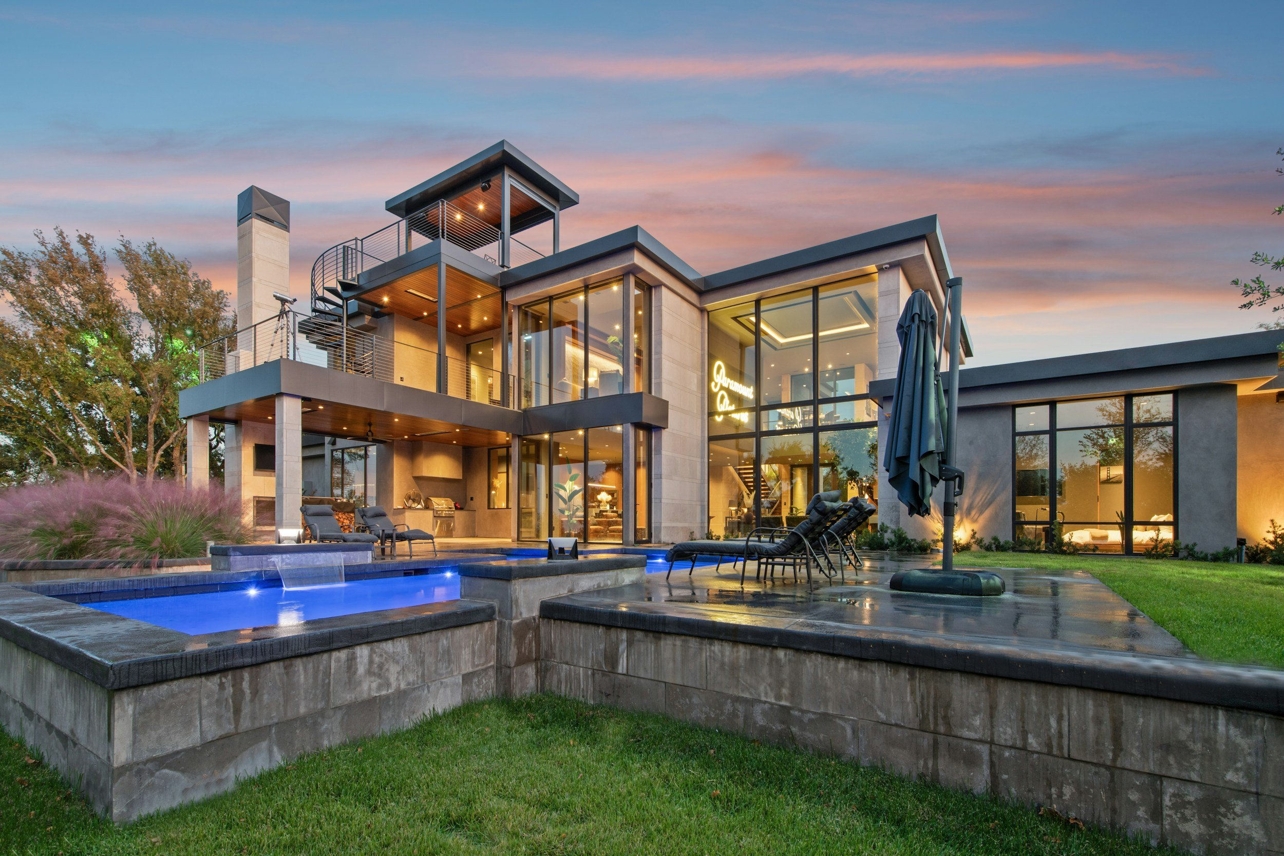 Modern house with large glass windows, outdoor pool, lounge chairs, and surrounding greenery under a sunset sky.