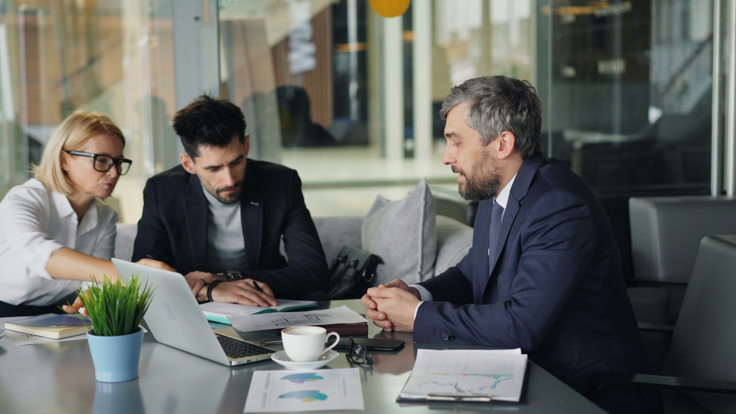 Three business professionals sitting at a table in a modern office, engaged in a serious discussion, with documents, a laptop, and a coffee cup in front of them.