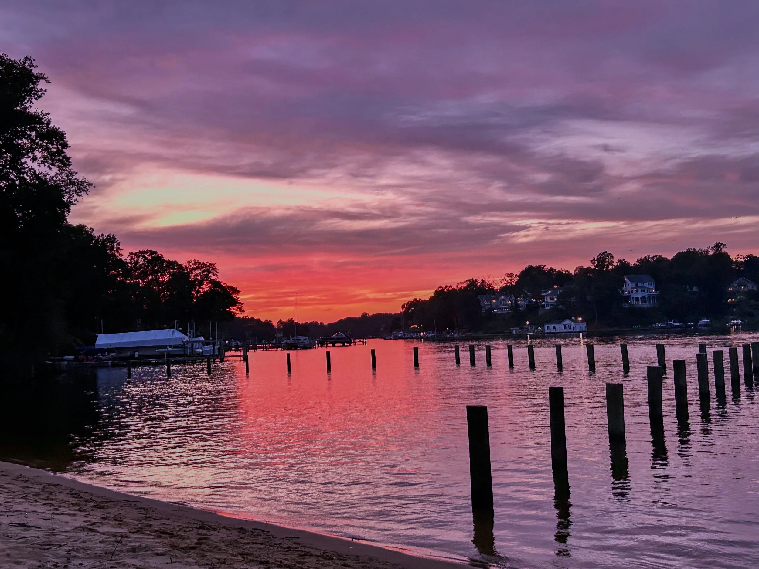 A colorful sunset over a calm river with silhouettes of trees and houses along the shoreline.