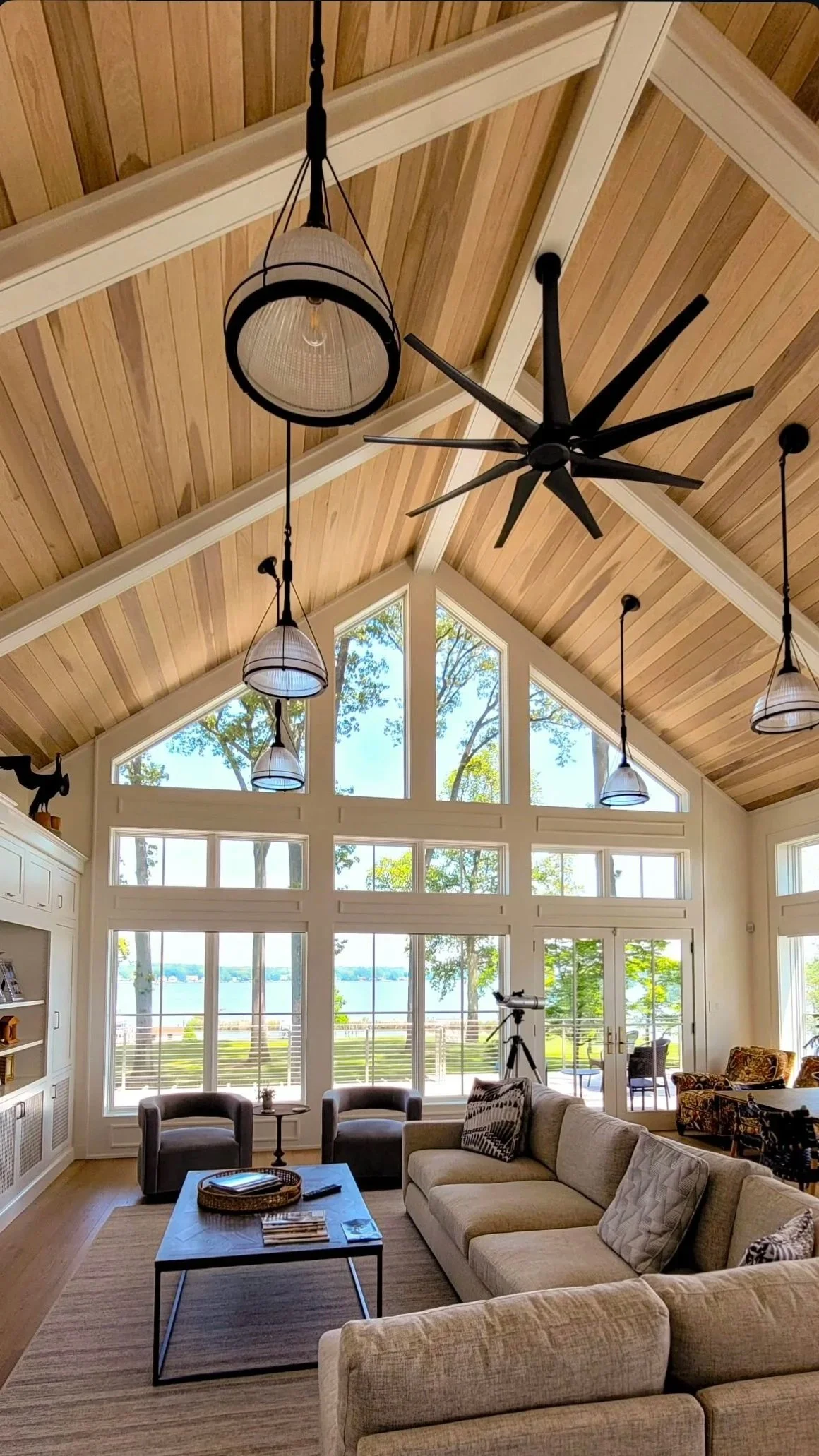 Living room with large windows showing a lake view, high vaulted wood ceiling with black ceiling fan and hanging light fixtures, beige sofas, and a telescope near the window.