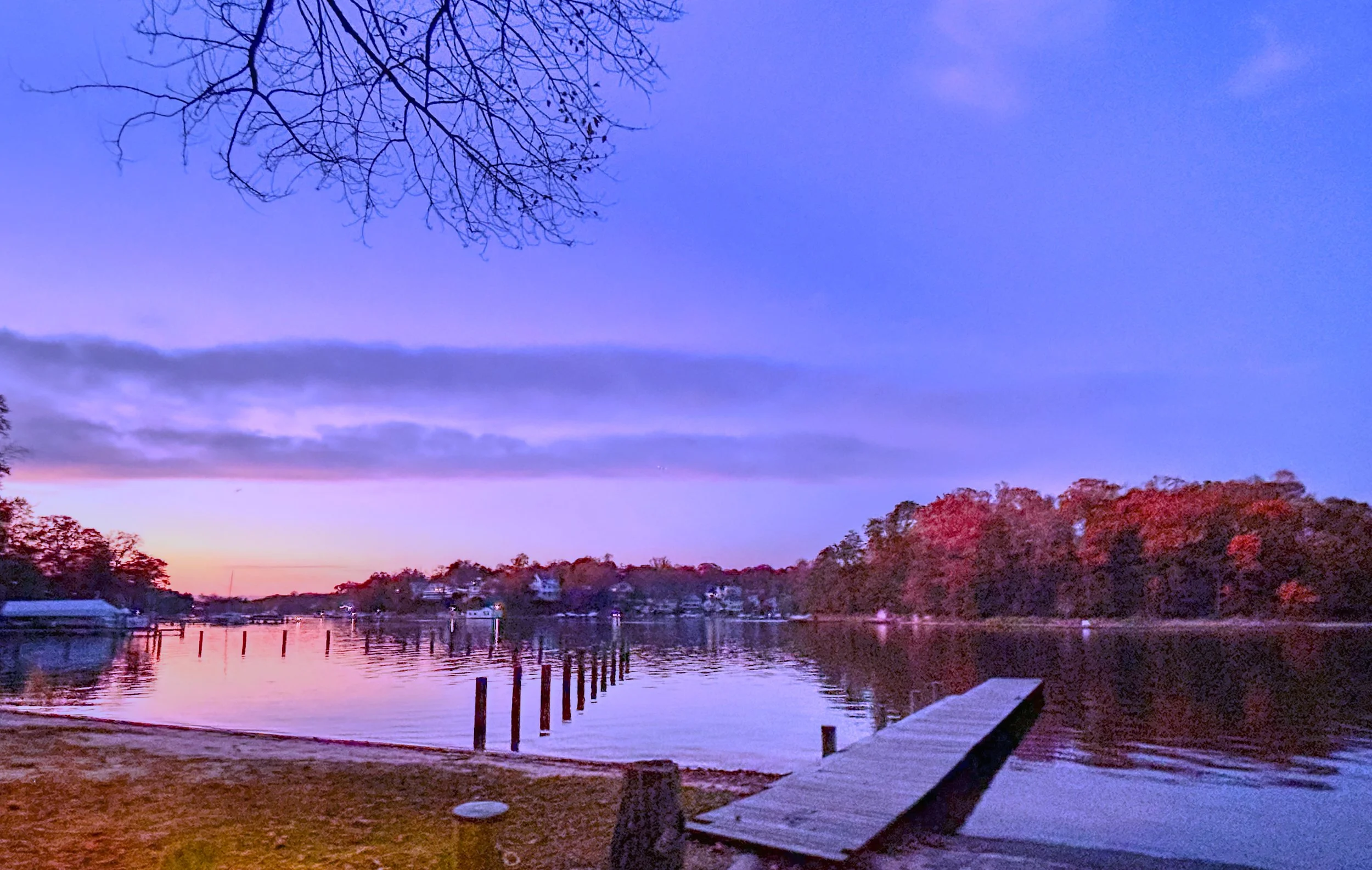 Serene lakeside scene at dusk with calm water reflecting the purple and pink hues of the sunset sky. Bare tree branches extend from the top left corner, and a dock with wooden posts extends into the water. Trees with red foliage line the horizon.