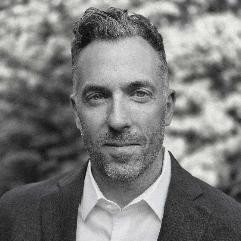 Un homme en costume avec une chemise blanche, regardant vers la caméra, en arrière-plan flou.