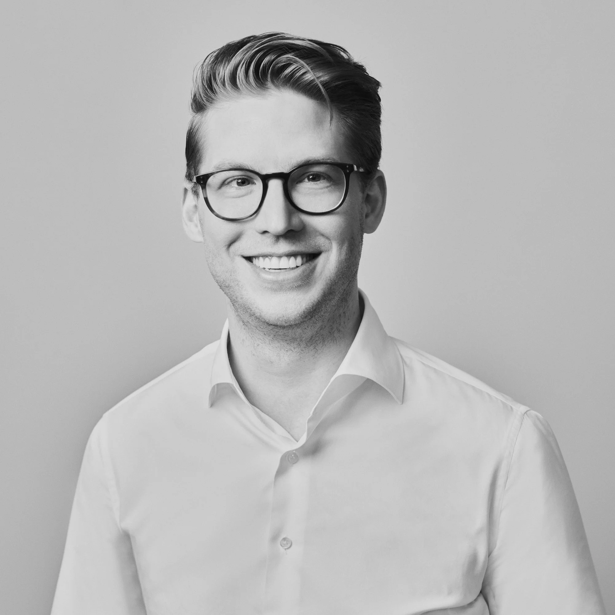 Portrait d'un homme souriant portant des lunettes et une chemise blanche, en noir et blanc.