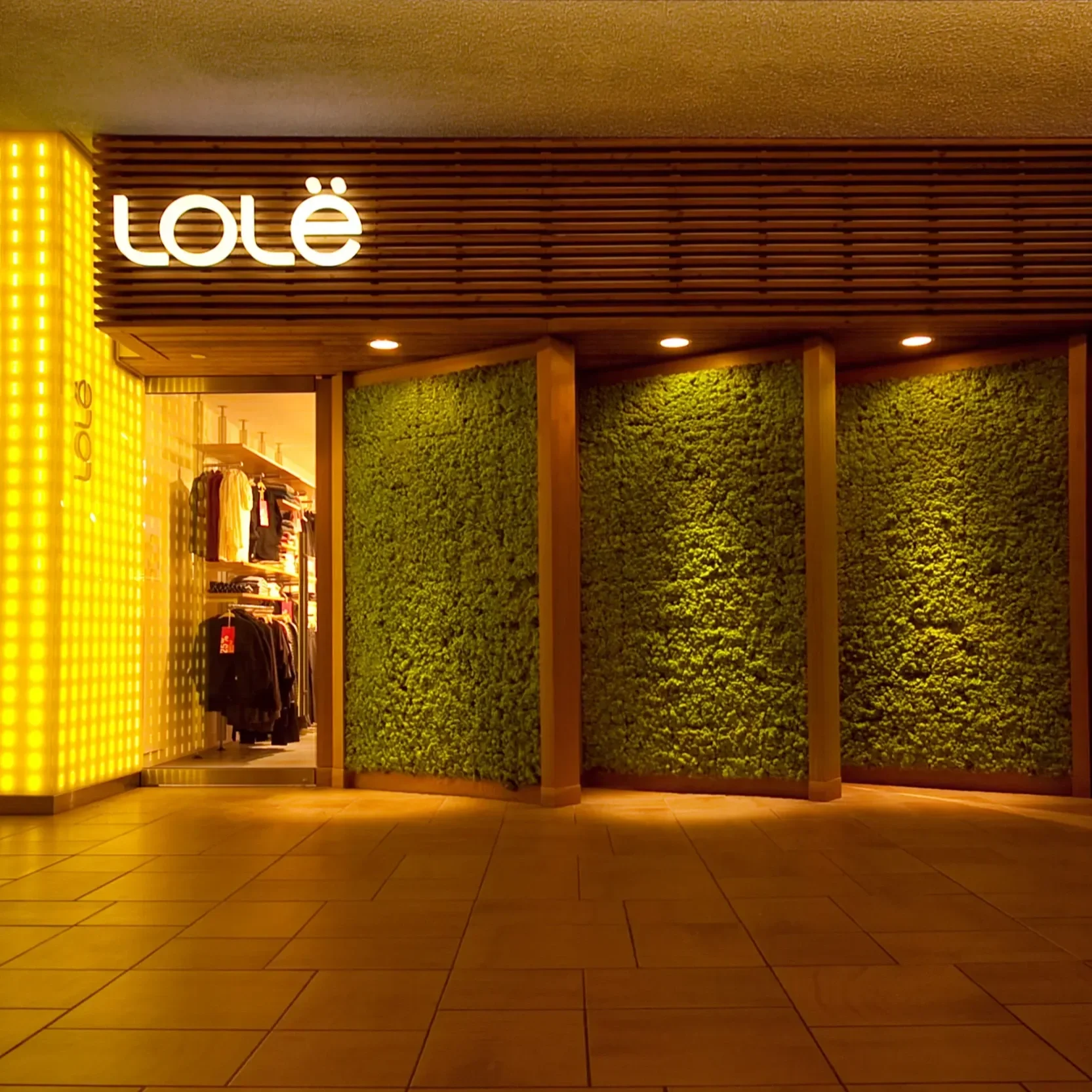 Vitrine d'une boutique avec une enseigne lumineuse 'Lolé', murs en bois et fausse végétation vert foncé, vêtements exposés à l'intérieur.