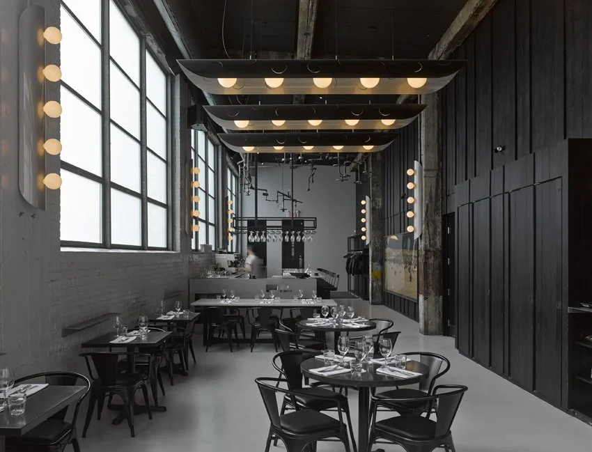 Intérieur d'un restaurant moderne avec grande fenêtre, lampes suspendues et tables dressées avec de la vaisselle et des verres.