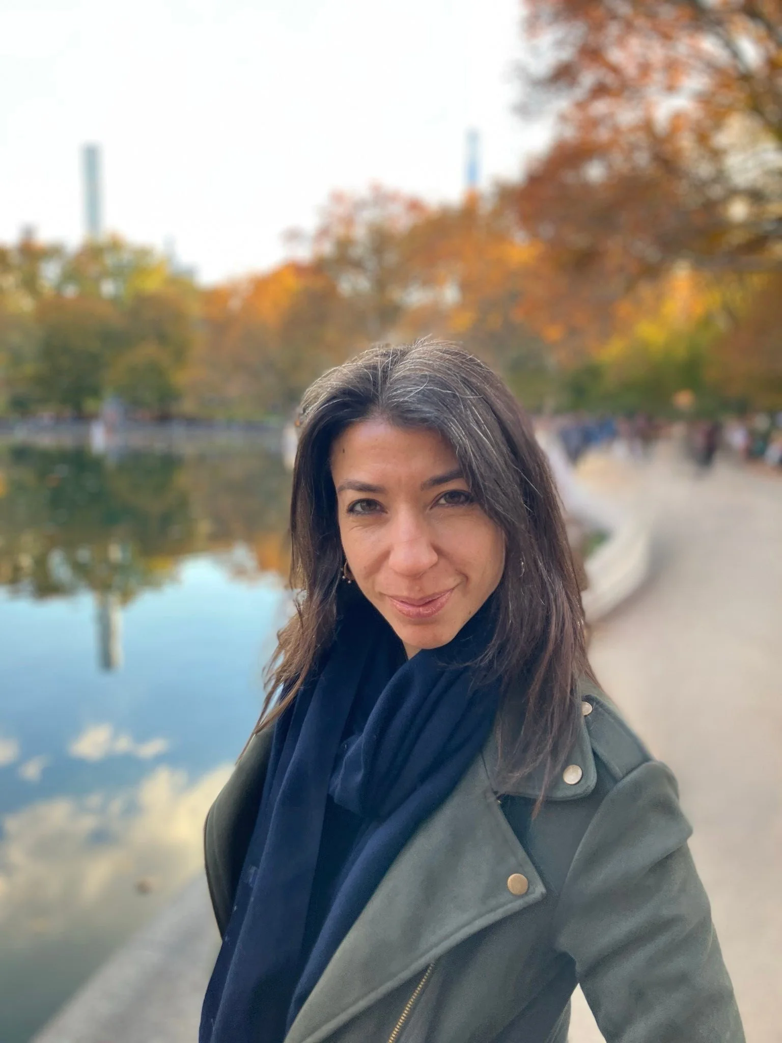A woman taking a selfie outdoors by a body of water with autumn trees in the background.