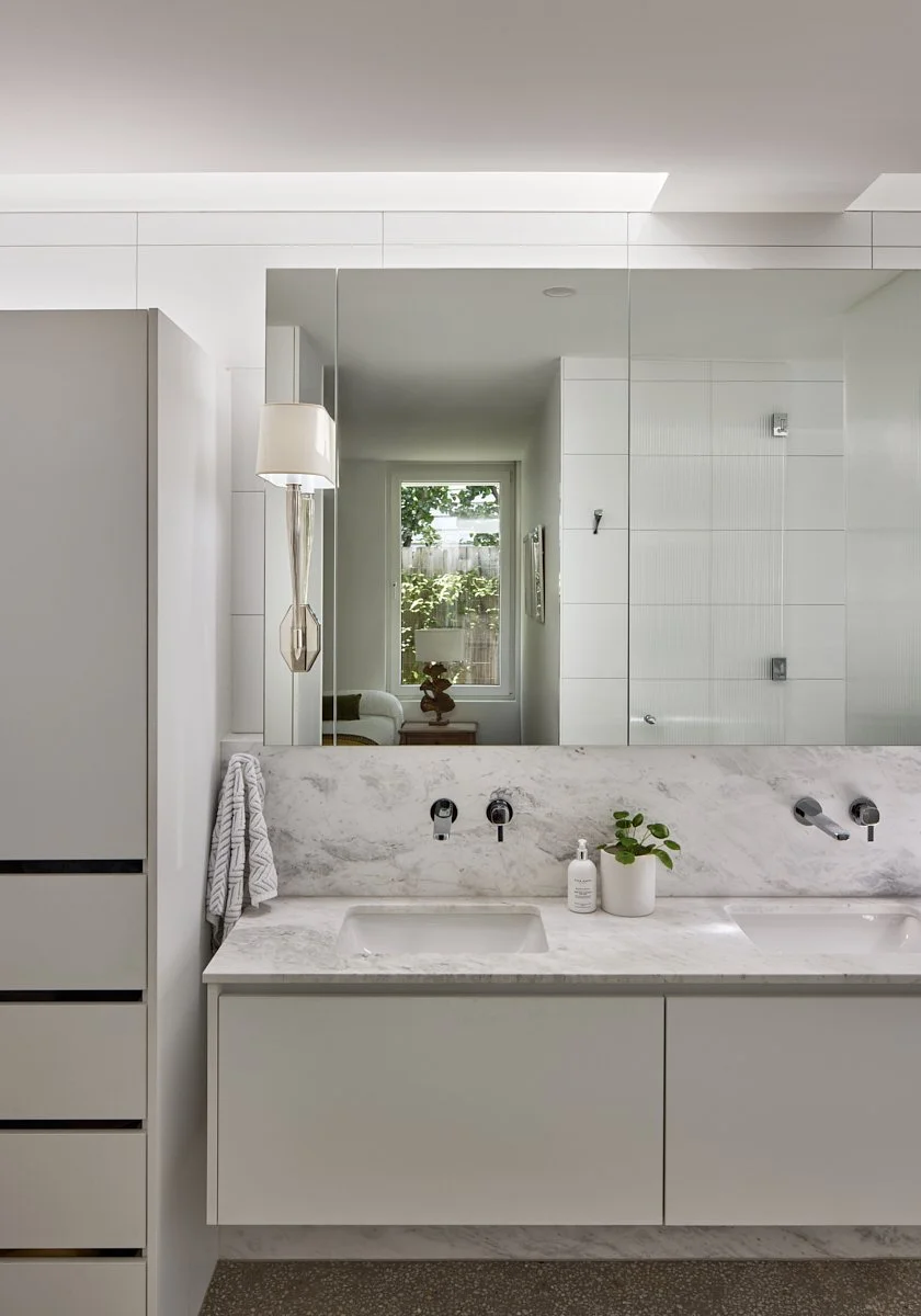 Modern bathroom with white marble countertop, dual sinks, and a large mirror reflecting a window and part of a sitting area with a lamp and plant.