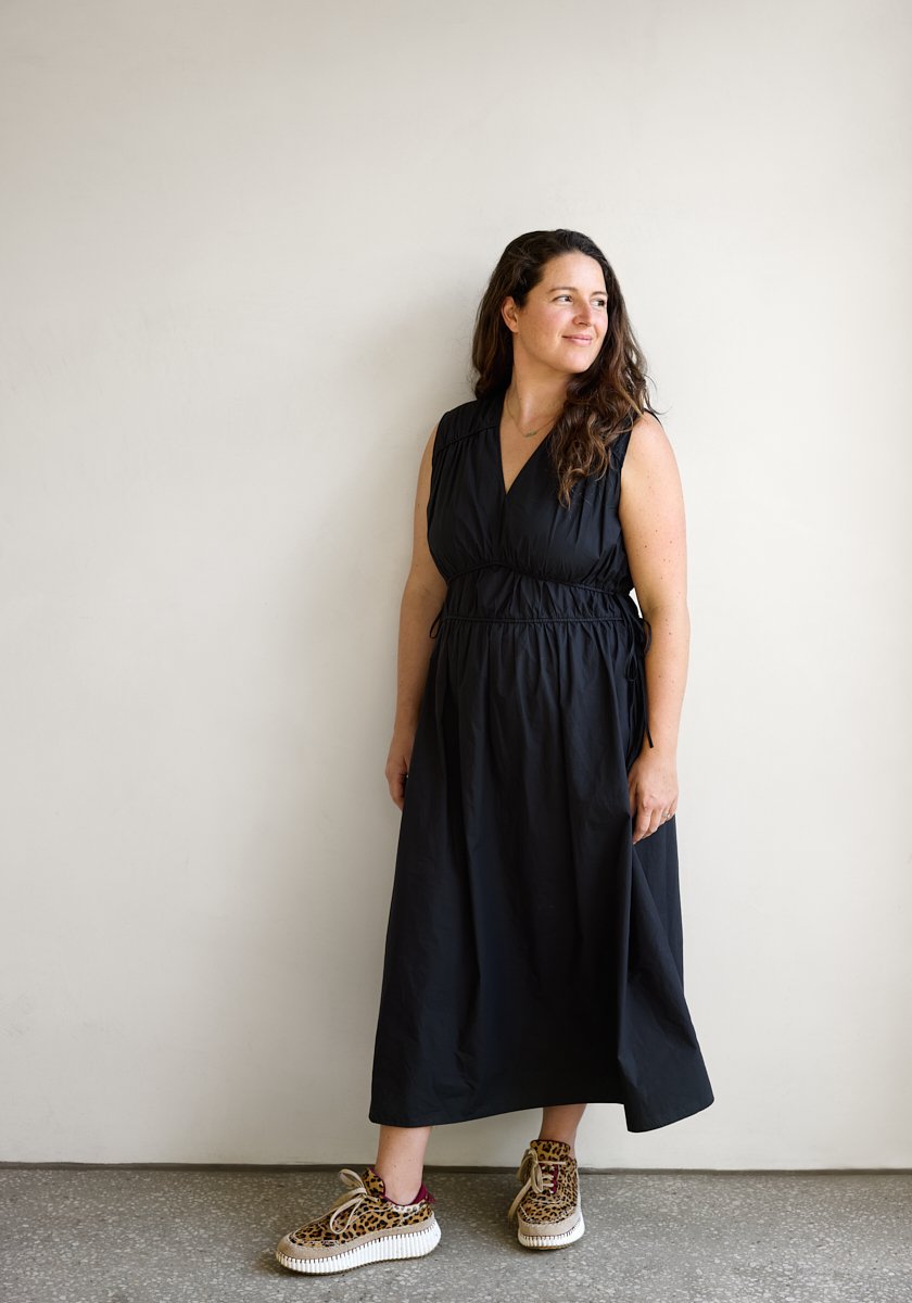 A woman standing against a plain white wall, wearing a sleeveless black maxi dress and leopard print sneakers, looking to the side with a slight smile.