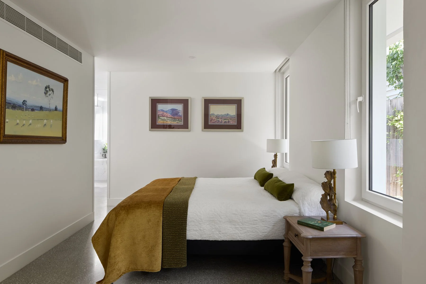 A bedroom with a white bed, an olive green bed runner, and matching pillows, beside a bedside table with a lamp and a book, large windows with natural light, and framed artwork on white walls.
