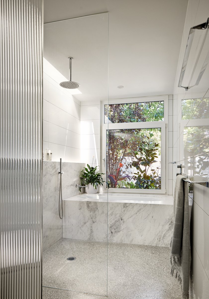 Modern bathroom with a walk-in shower, marble bathtub, large window revealing greenery outside, white tiled walls, plants, and a gray towel hanging on a rack.