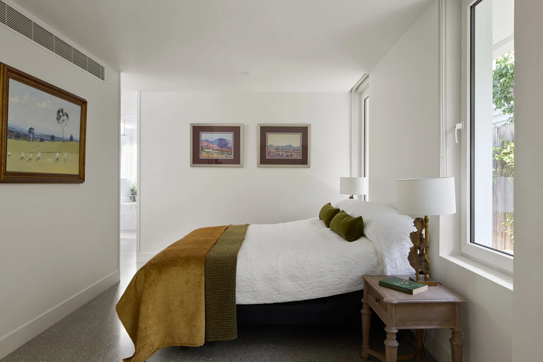 A cozy bedroom with a white bedspread, green pillows, and a brown blanket. Natural light comes through a large window. Wall art includes landscape paintings. A small wooden bedside table with a book and a brass lamp with a white shade. The room featu