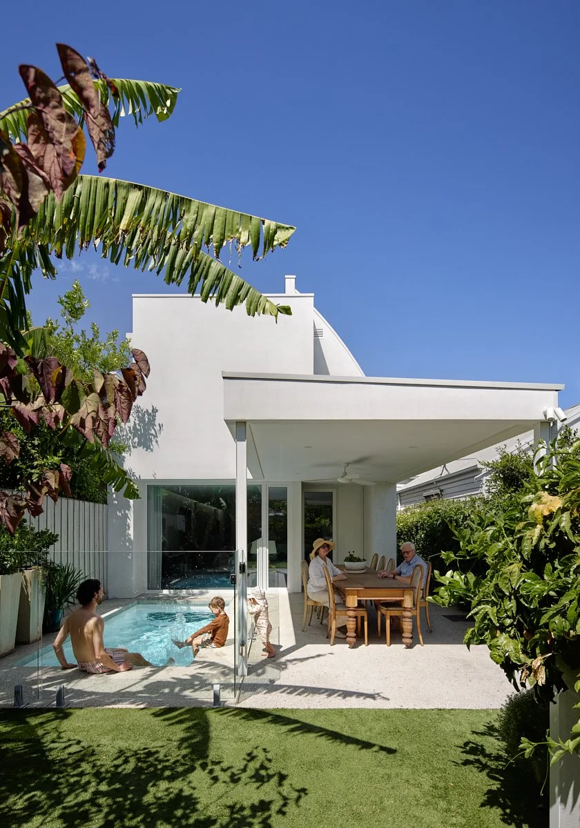 People relaxing in a backyard with a pool, dining area, and lush green plants, under a clear blue sky.