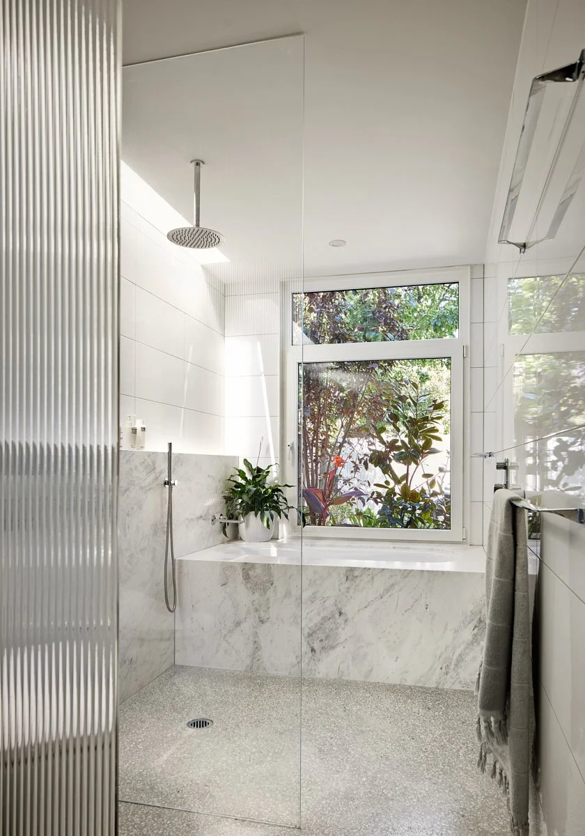 Modern bathroom with a large window overlooking greenery, marble bathtub, plant, and shower area with a rain shower head.