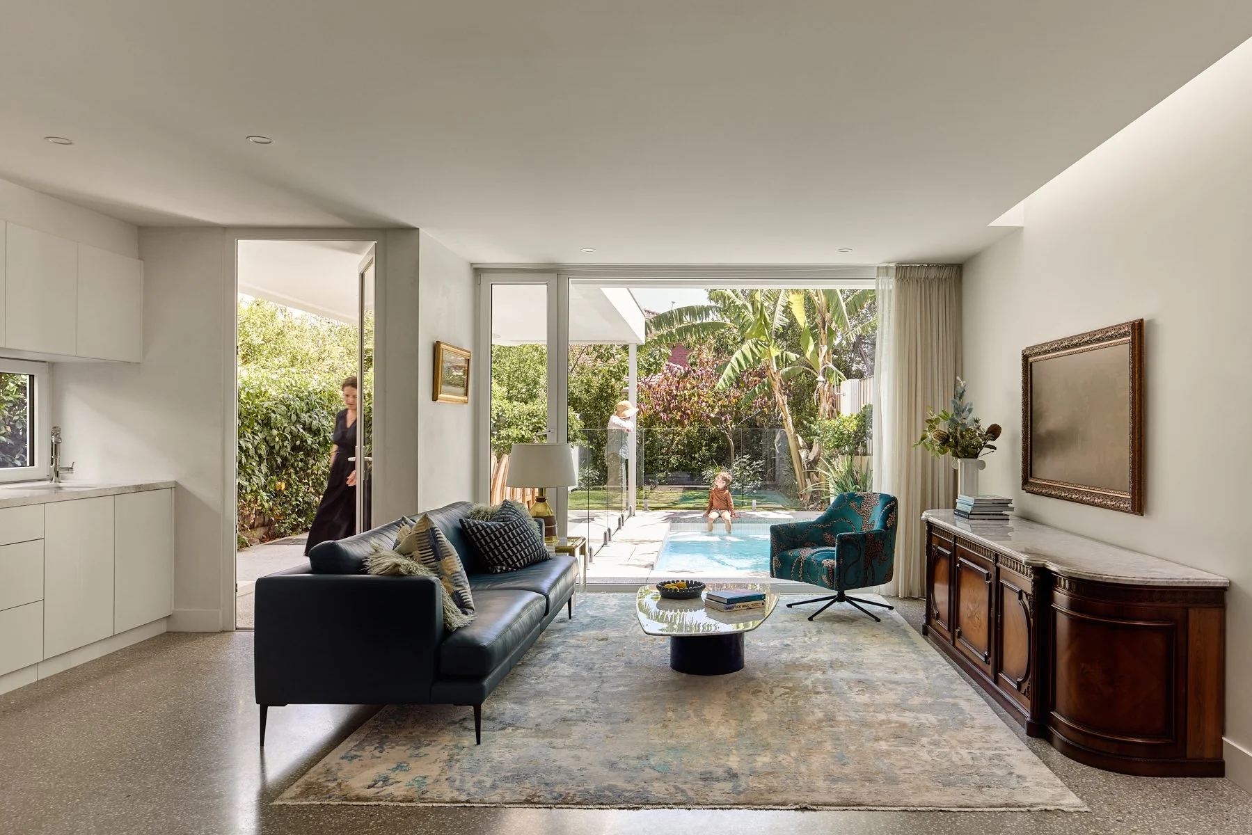 Living room with large glass sliding door opening to a backyard pool and garden, with people visible outside. Interior contains a black sofa, colorful armchair, marble-topped sideboard with a vase of flowers, framed artwork, and a coffee table on a p