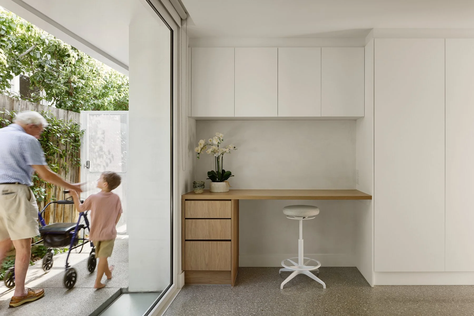 Minimalist indoor workspace with a built-in desk, white cabinets, a potted orchid, and a white adjustable stool. Outside the open glass door, an elderly man and a young girl are playing with a walker on a patio surrounded by greenery.