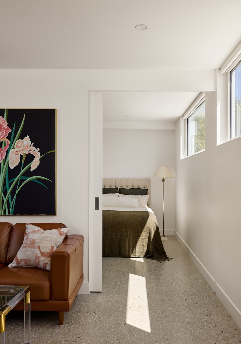 View of a modern living room with a leather sofa, a decorative pillow, a large floral painting, and a partial view of a bedroom with a bed and floor lamp, sunlight streaming through windows.