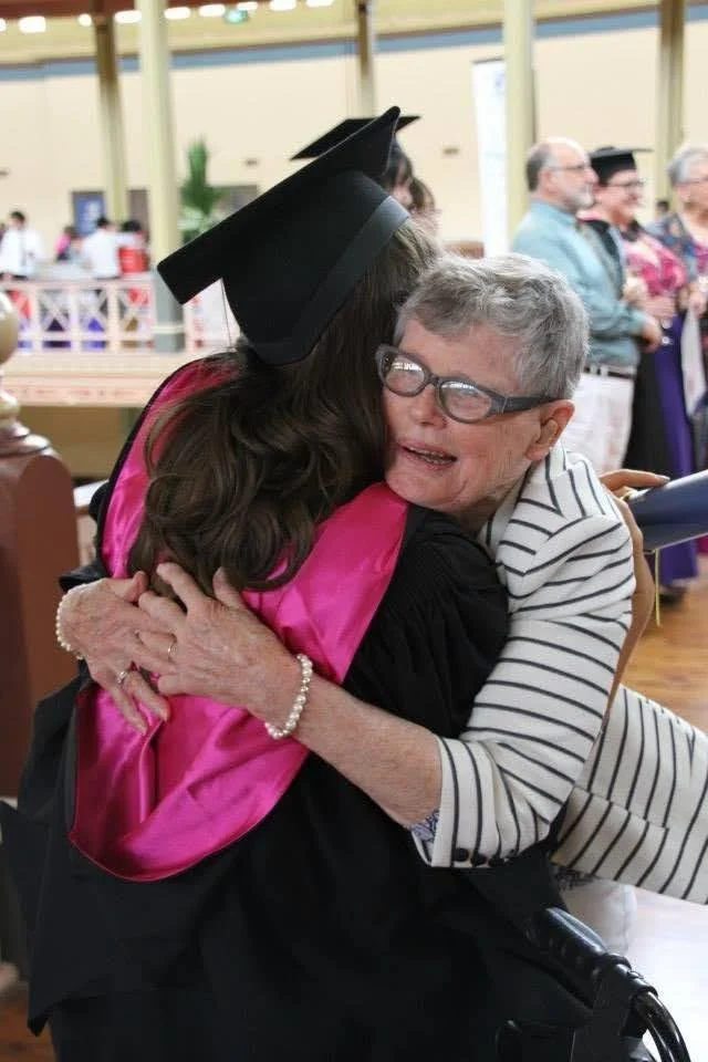 A young woman in a cap and gown hugs an elderly woman at a graduation ceremony.