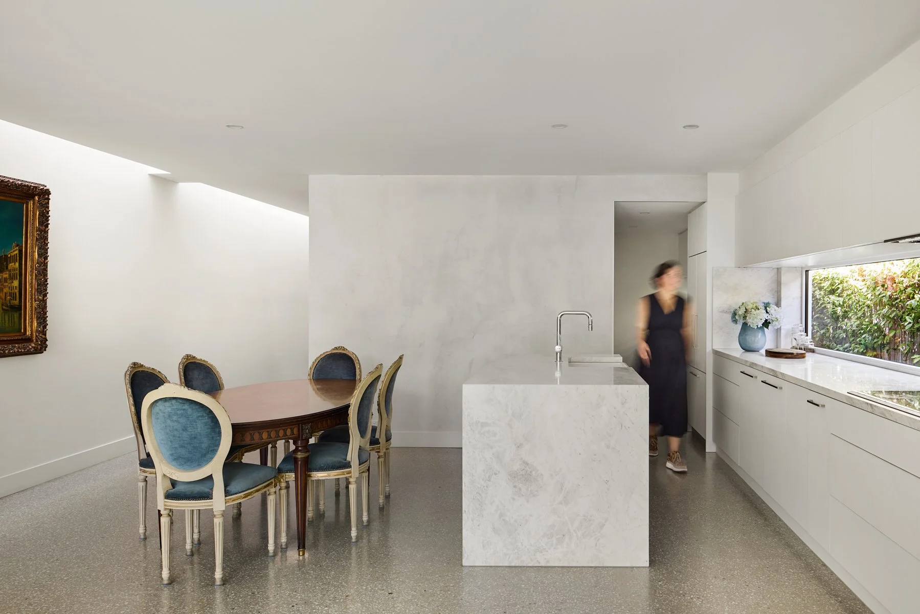 Modern kitchen with white marble island, white cabinetry, window with greenery outside, and a round dining table with blue upholstered chairs. A woman is walking past in the background.