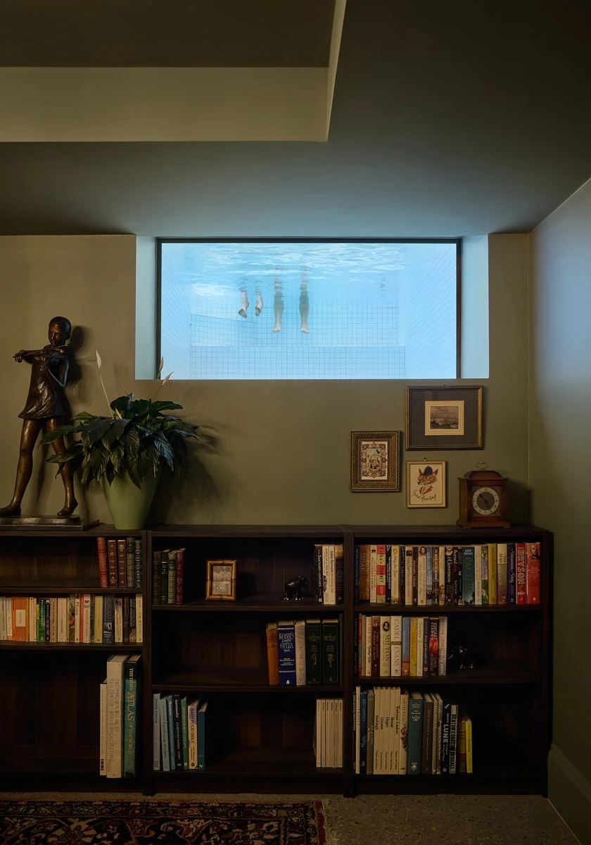 Living room with bookshelf, framed pictures, a clock, a large potted plant, and a window showing people's legs underwater.