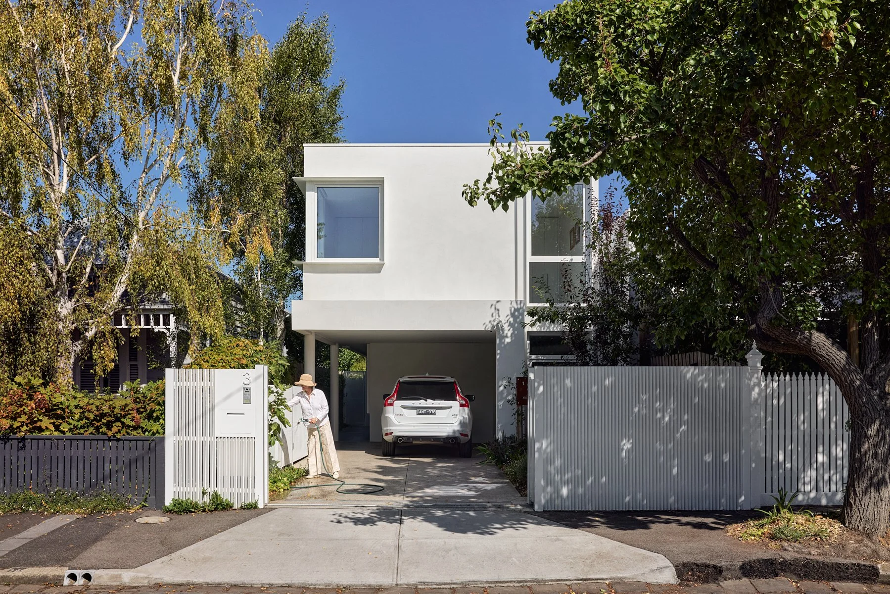Modern white residential house with a driveway and a car parked underneath the second story. A person wearing a hat and light-colored clothing is watering plants outside the gate. There are trees and a blue sky in the background.