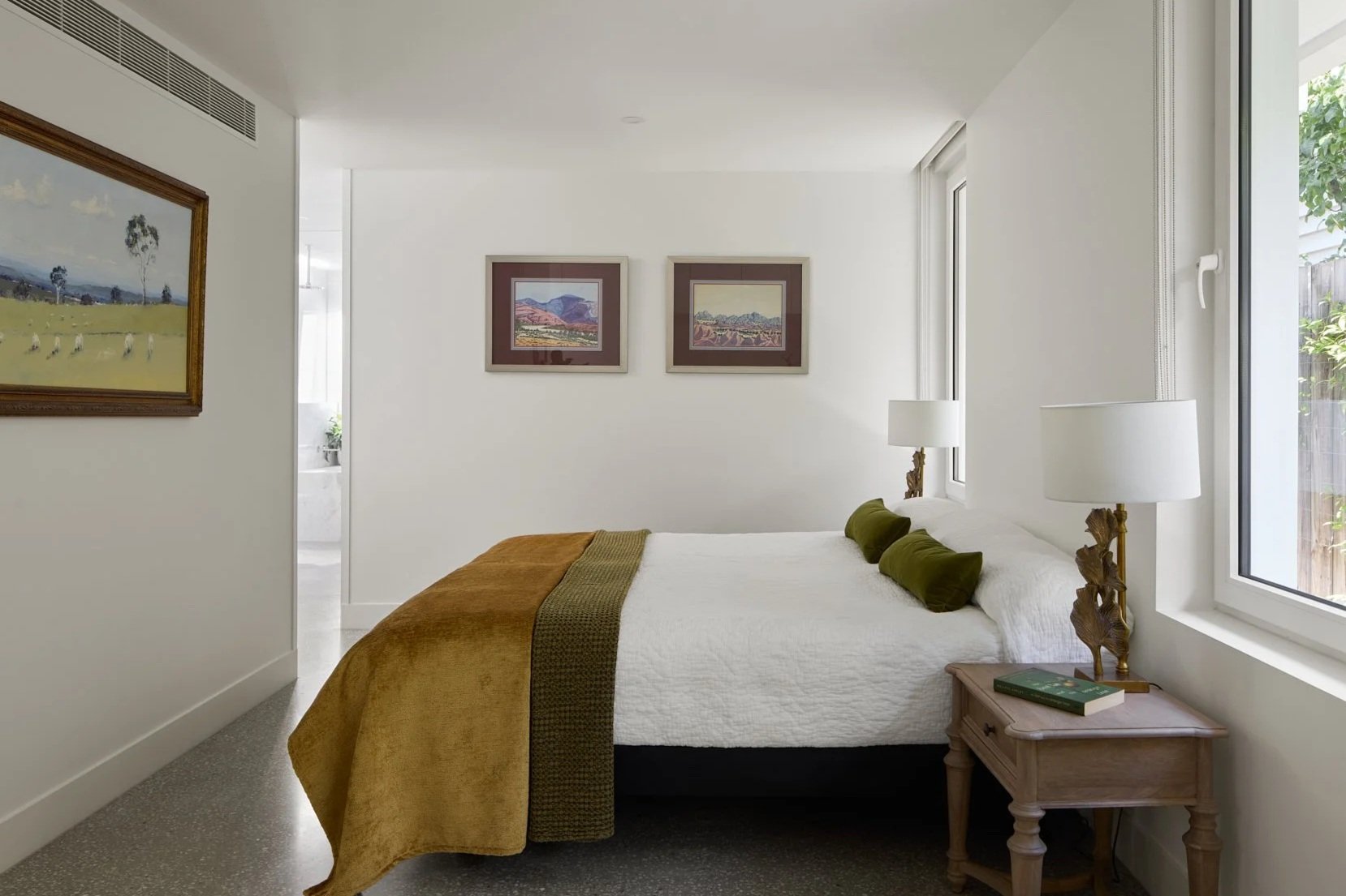 A bedroom with white walls, featuring a bed with white bedding, green pillows, and a brown throw blanket, flanked by two bedside tables with lamps. There are framed landscape artwork on the walls, a window with greenery outside, and a partial view into an adjacent bathroom.