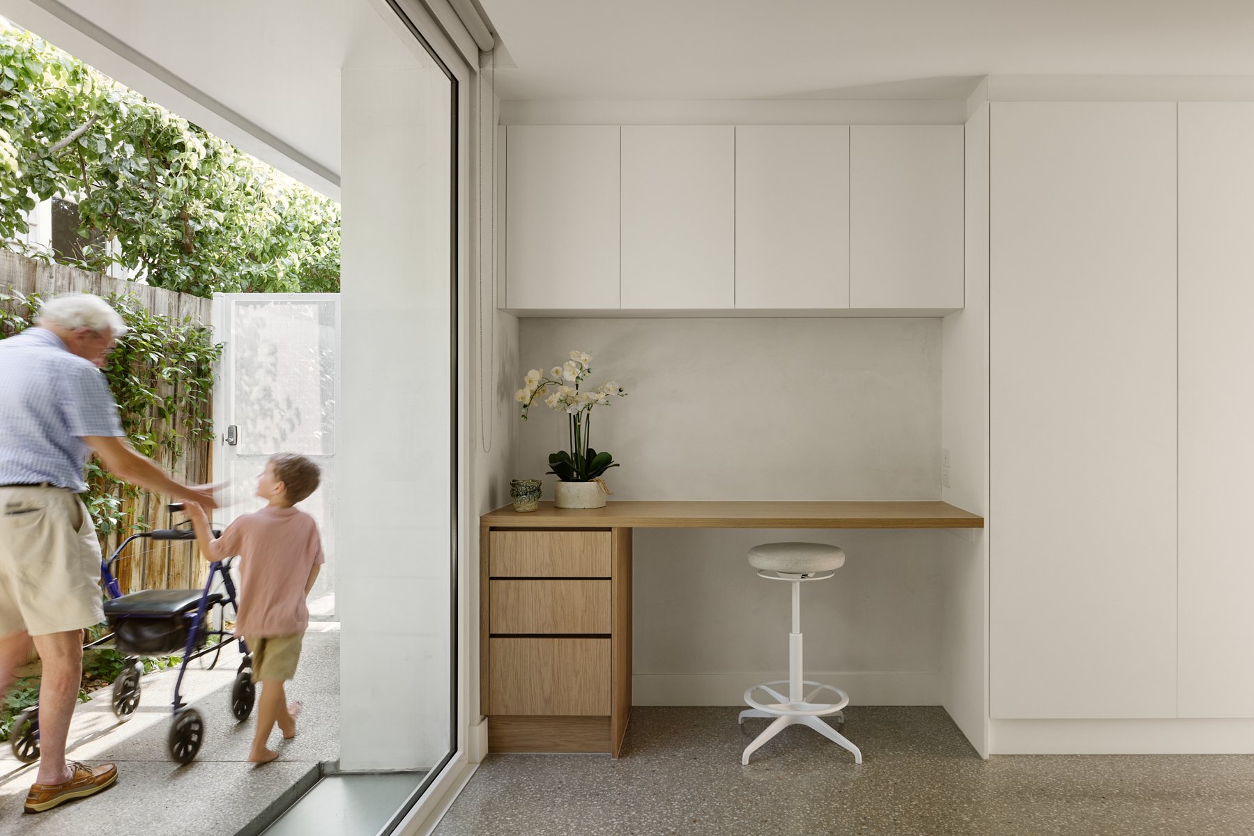 Interior of a minimalist room with white cabinets, a wooden desk, a white swivel stool, and a potted orchid plant. Outside, an elderly man and a young boy are seen through a sliding glass door, playing outside near a wooden fence and greenery.
