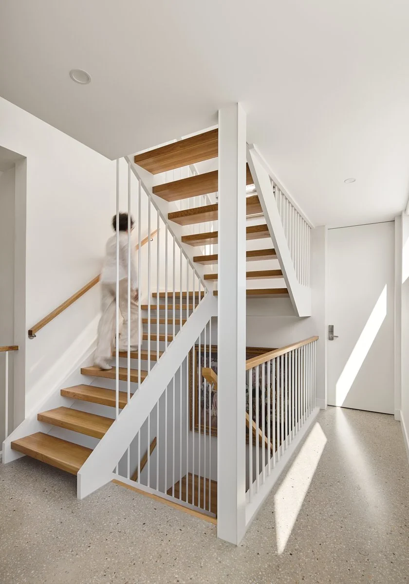 Interior view of a staircase with wooden steps, white balustrades, and a half-landing, with a woman ascending and sunlight streaming in.