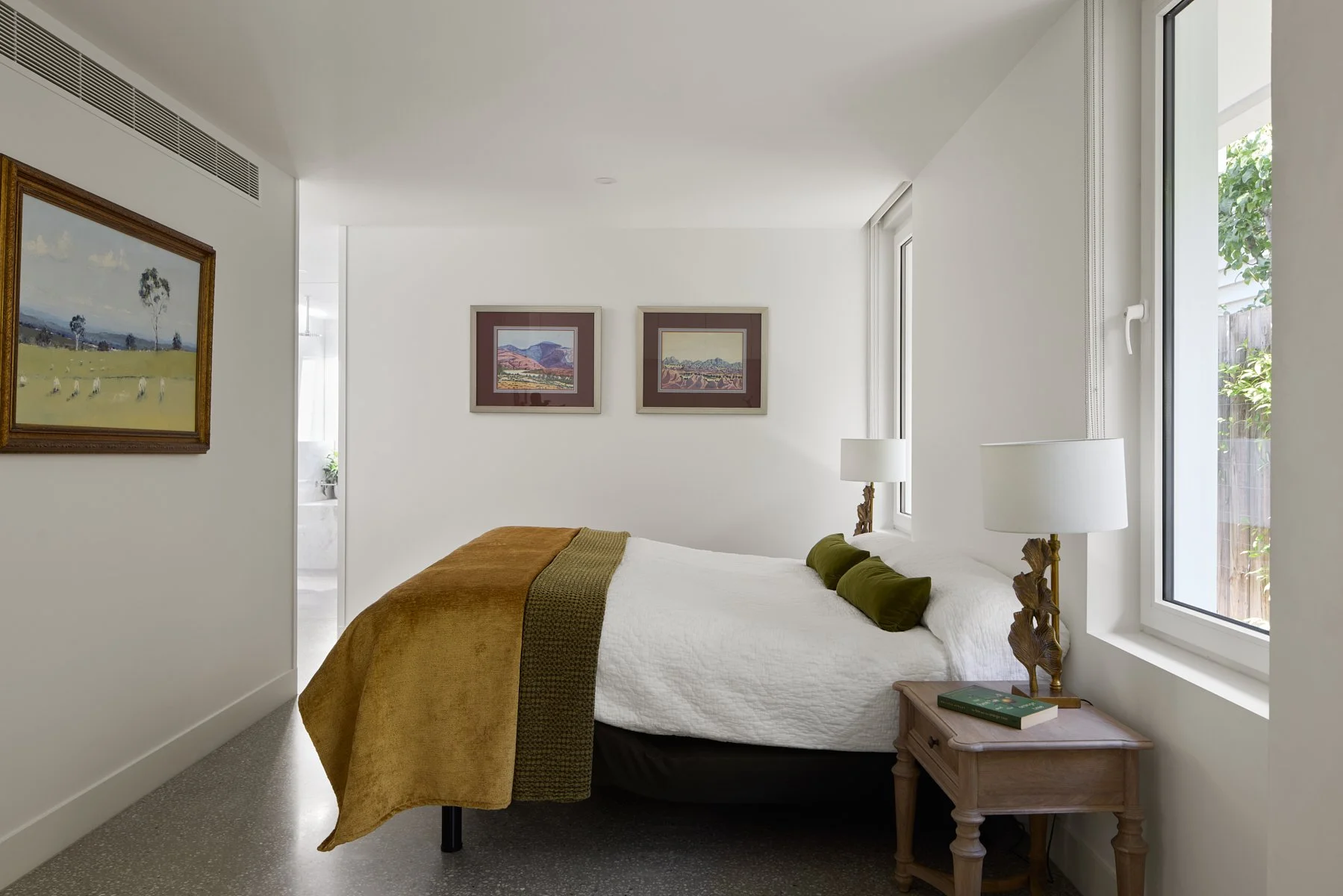 A bedroom with a bed covered in white and brown bedding, green pillows, and two table lamps on wooden nightstands by windows, white walls decorated with framed landscape art, and a view of a garden outside.