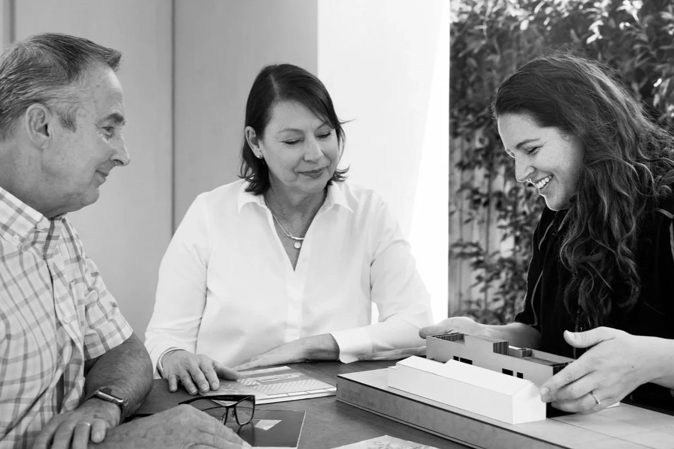 Three people sit at a table, discussing and examining a scale model of a building or house, in an indoor setting with plants in the background.