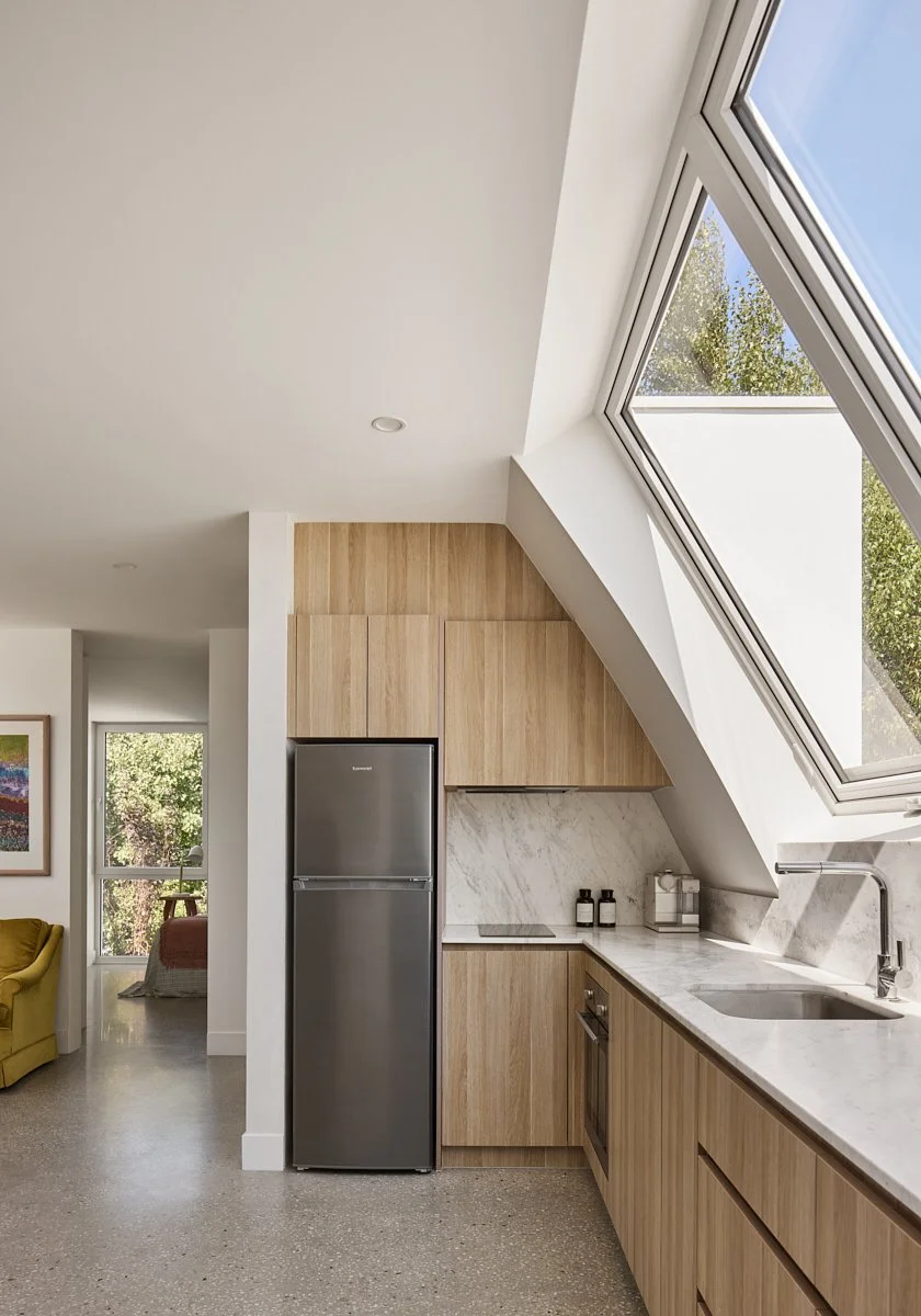 Modern kitchen with light wooden cabinets, stainless steel refrigerator, marble countertops, and a large skylight window.