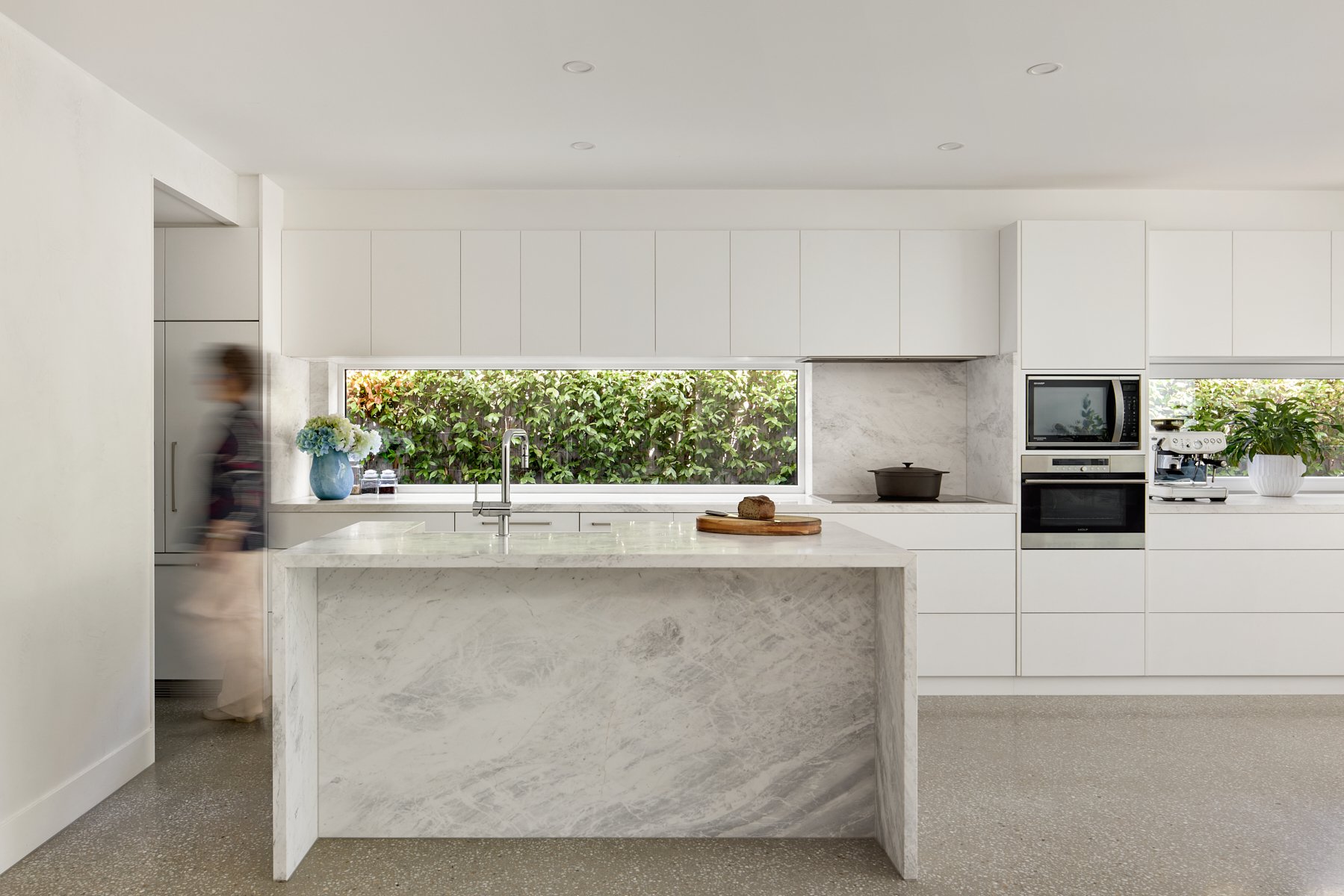 Modern white kitchen with a breakfast bar, built-in appliances, large window with greenery outside, and a person walking past.