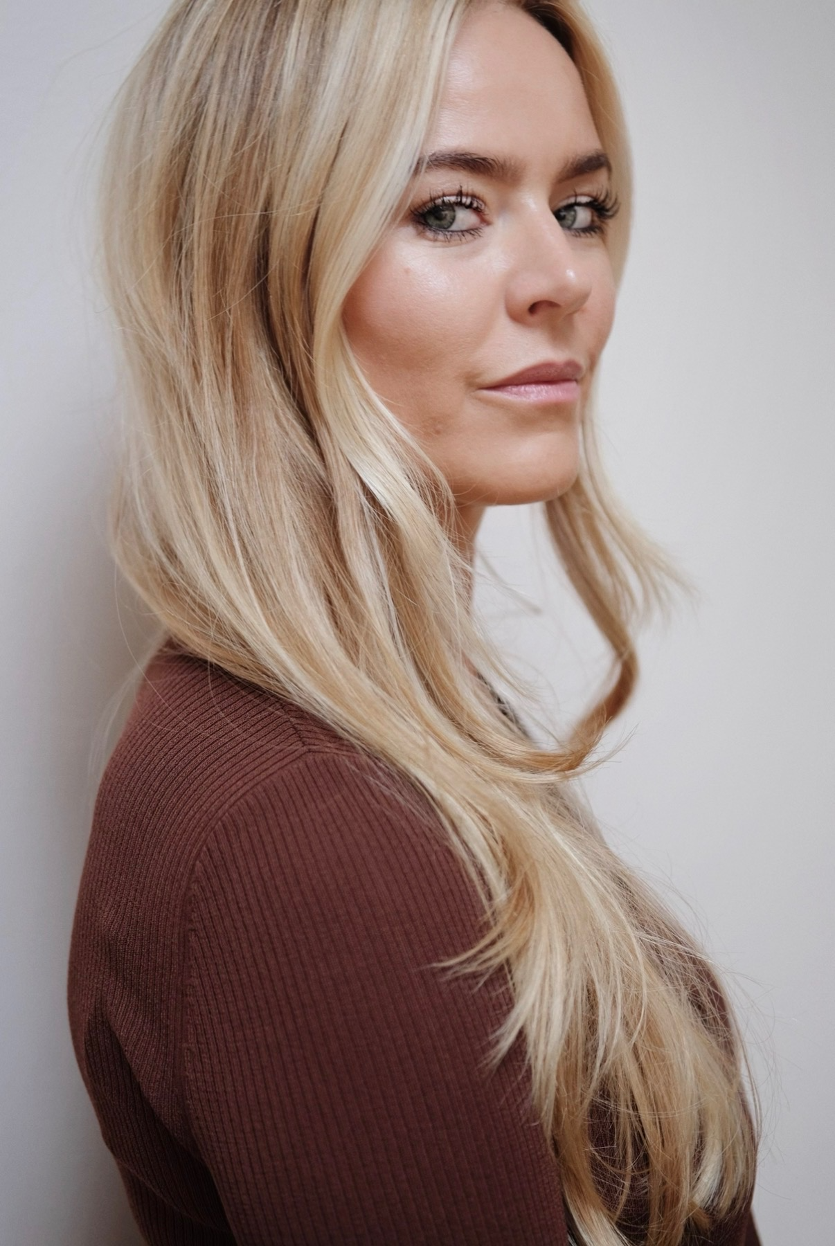A woman with long blonde hair and light makeup looking over her shoulder, wearing a brown long-sleeve top, standing against a light-colored plain background.