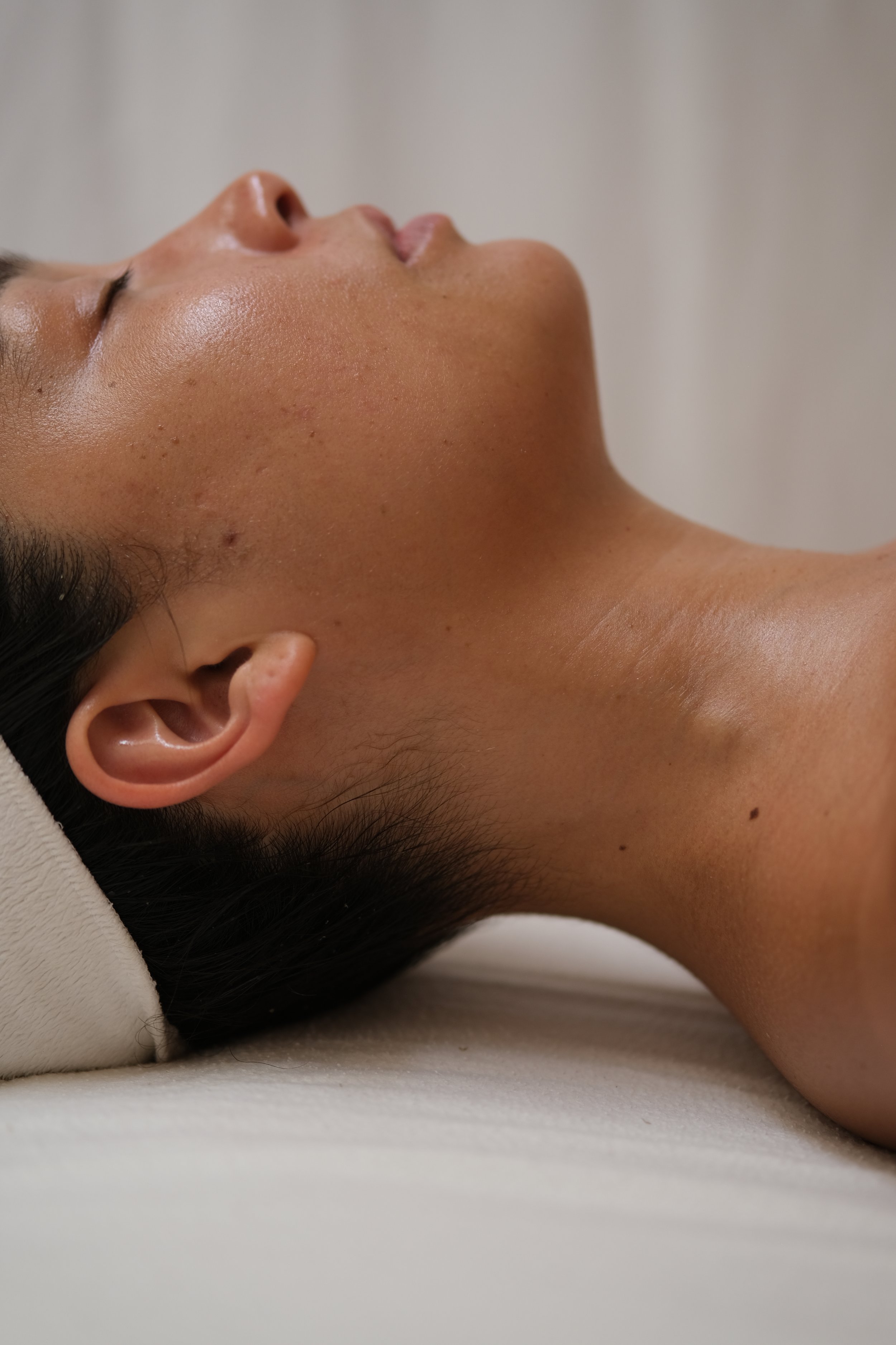Close-up side view of a person with eyes closed, lying down with head tilted back, skin appears smooth and glowing.