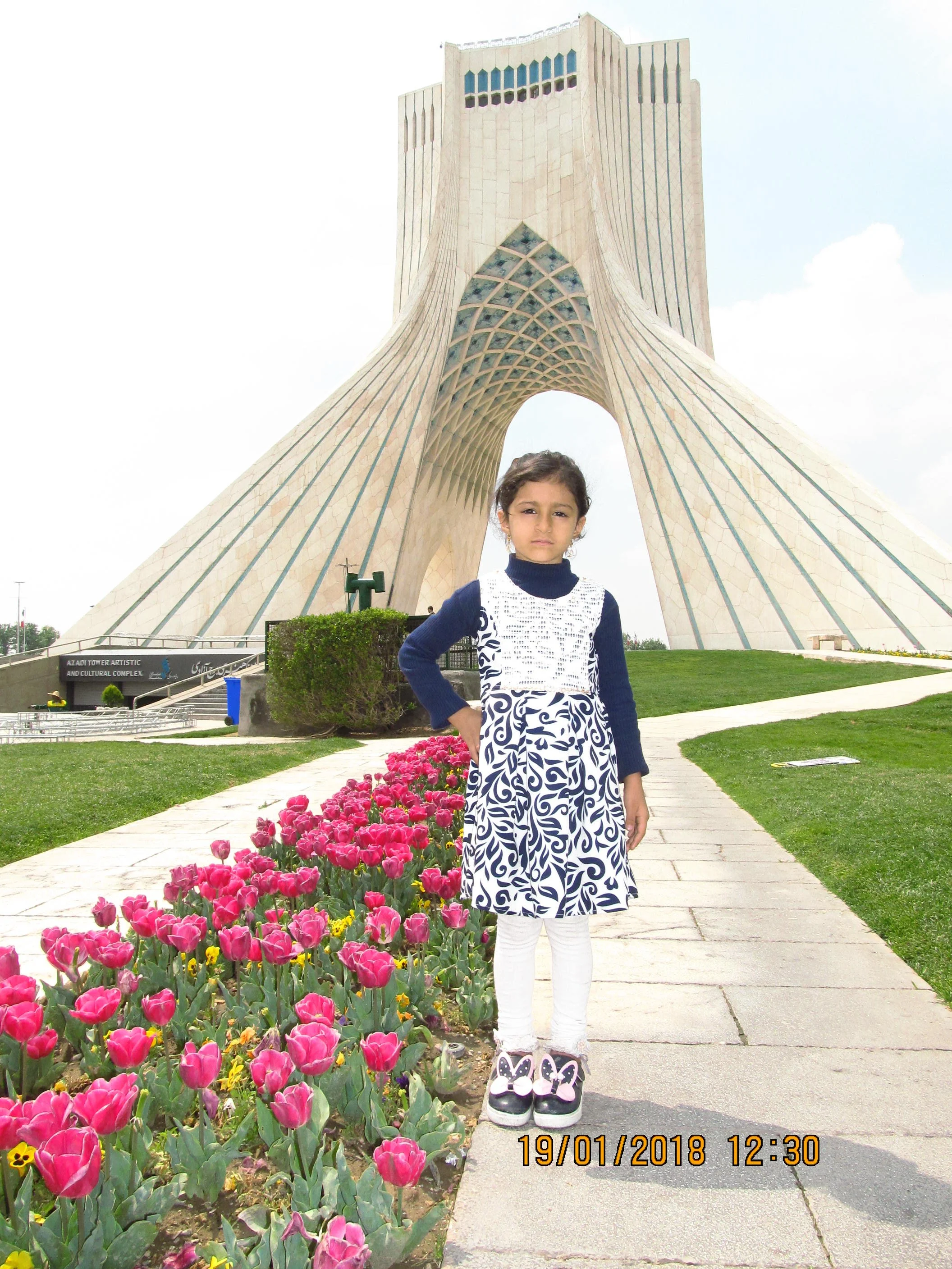 A young girl standing on a pathway in front of the Azadi Tower with pink tulips and green grass on either side.