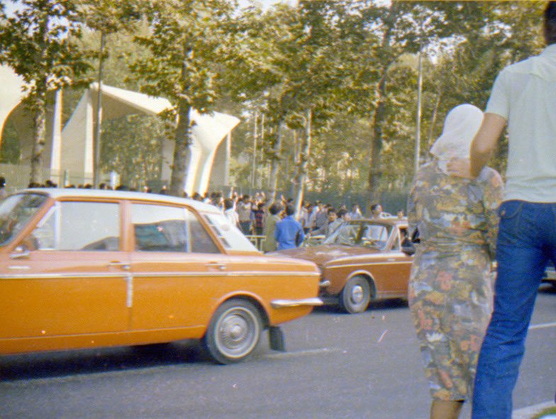 People gather outdoors with vintage cars in a park-like setting with trees and a modern white tent structure.