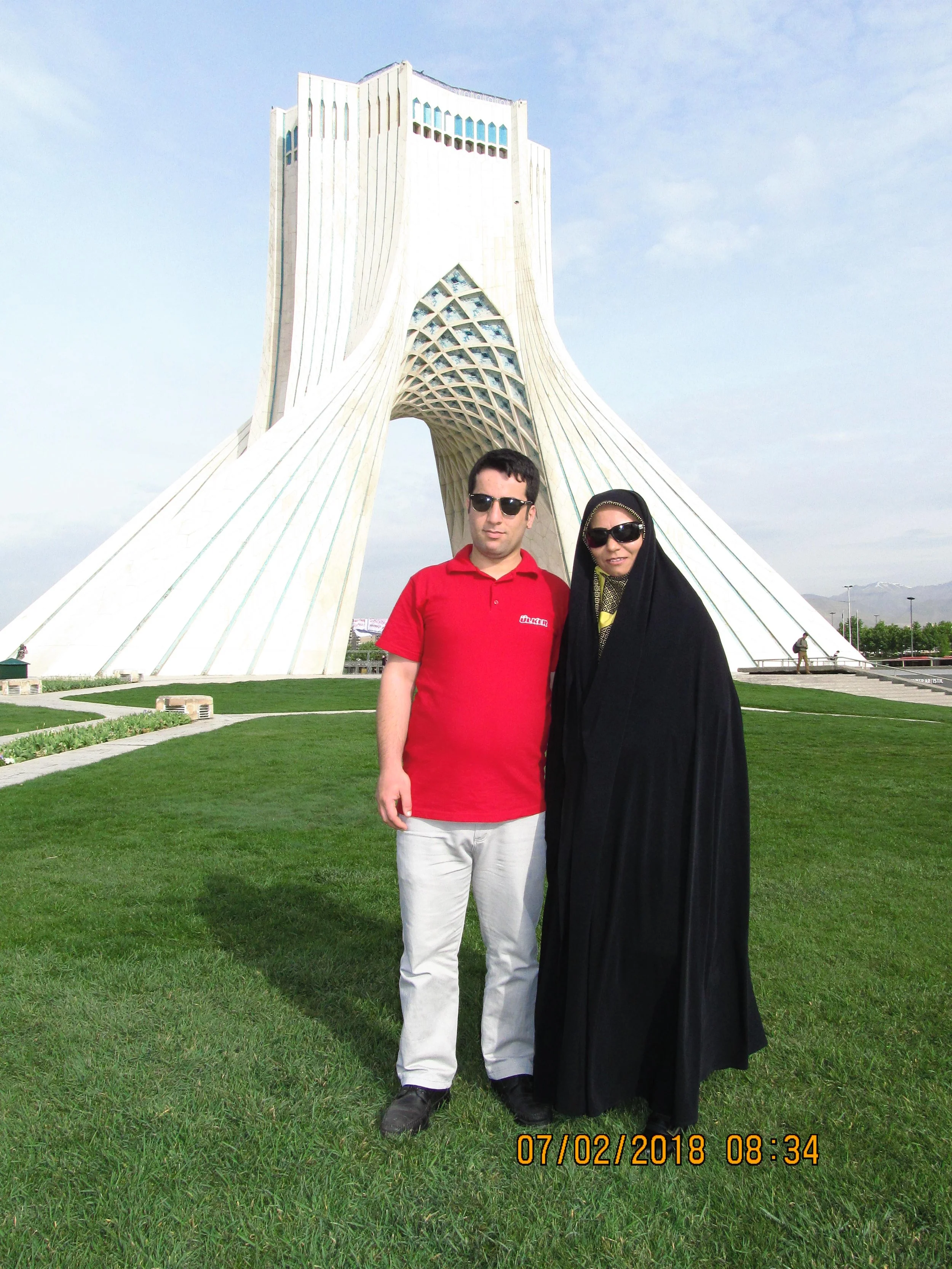 A man in a red shirt and a woman in black traditional clothing stand together on a green lawn in front of the Azadi Tower in Tehran, Iran, on a partly cloudy day.