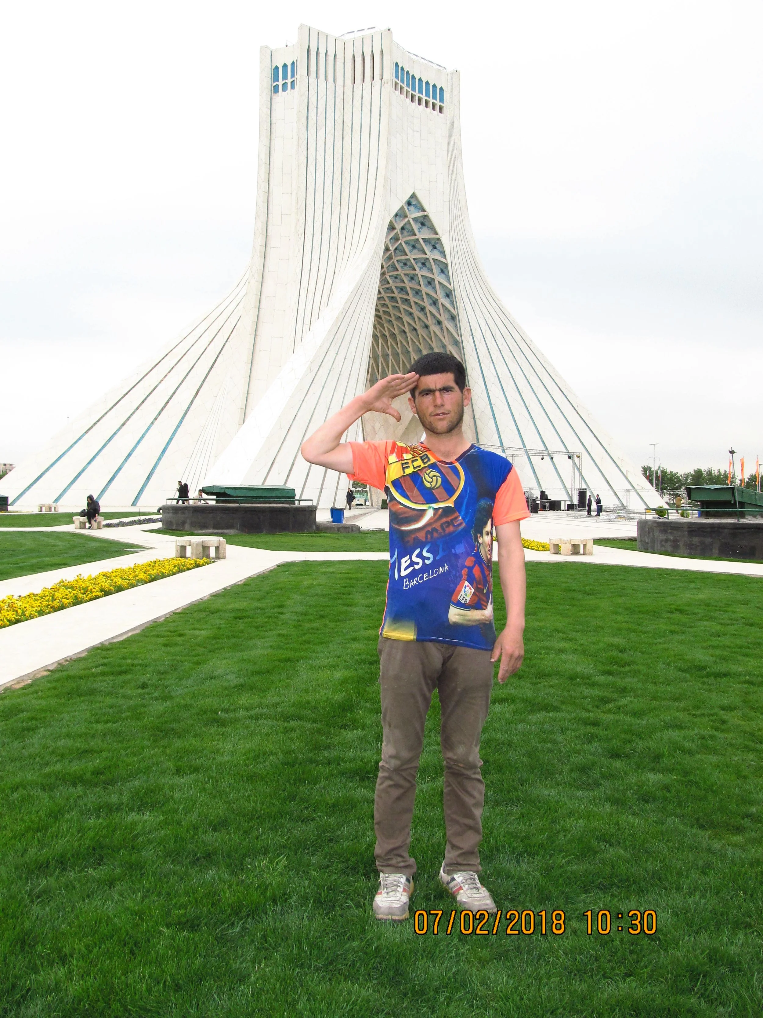 Young man saluting in front of a modern white architectural building, standing on green grass, with a yellow date and time stamp at the bottom.