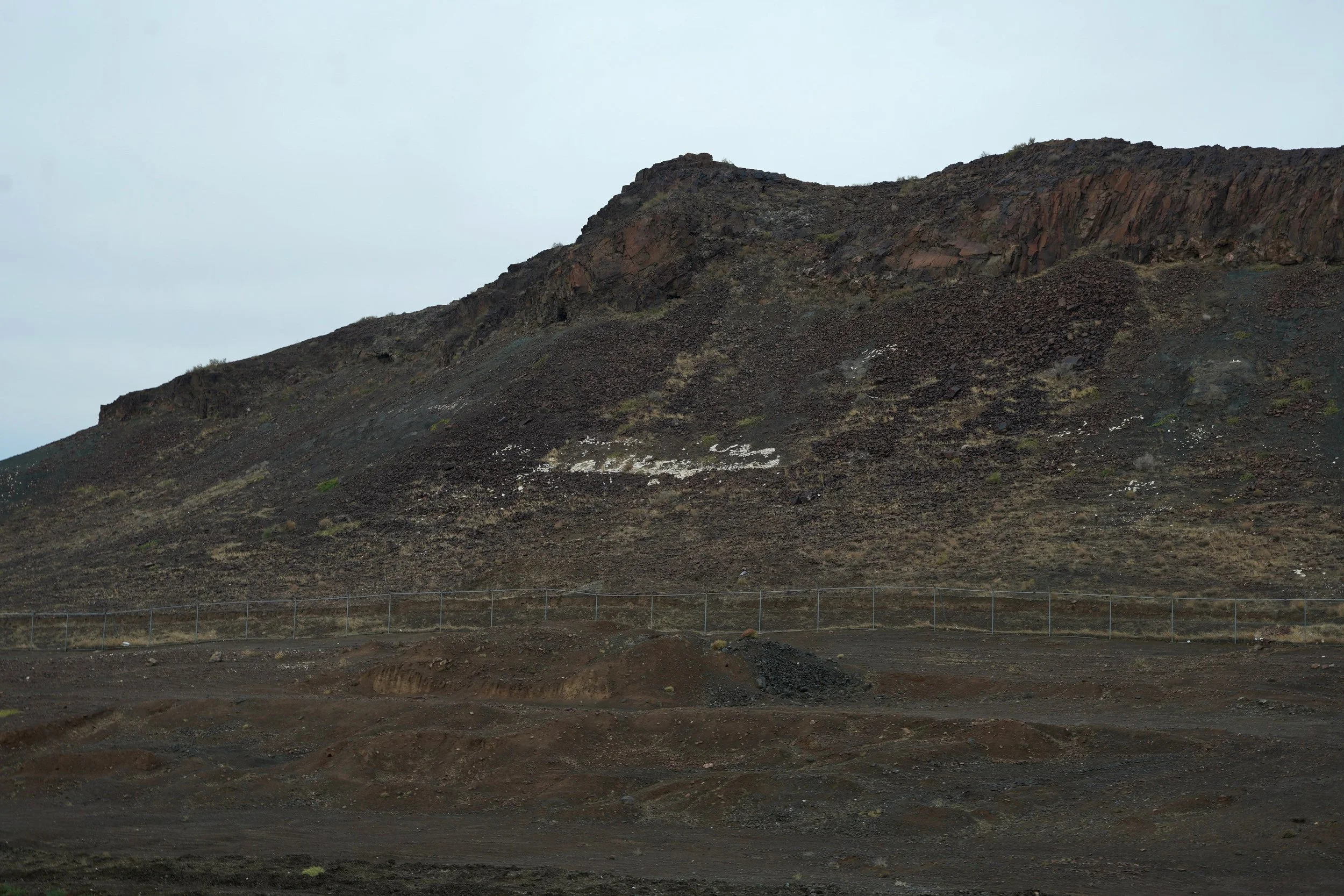 A barren mountain landscape with dark, rugged terrain and a fence running across the middle of the image, under an overcast sky.