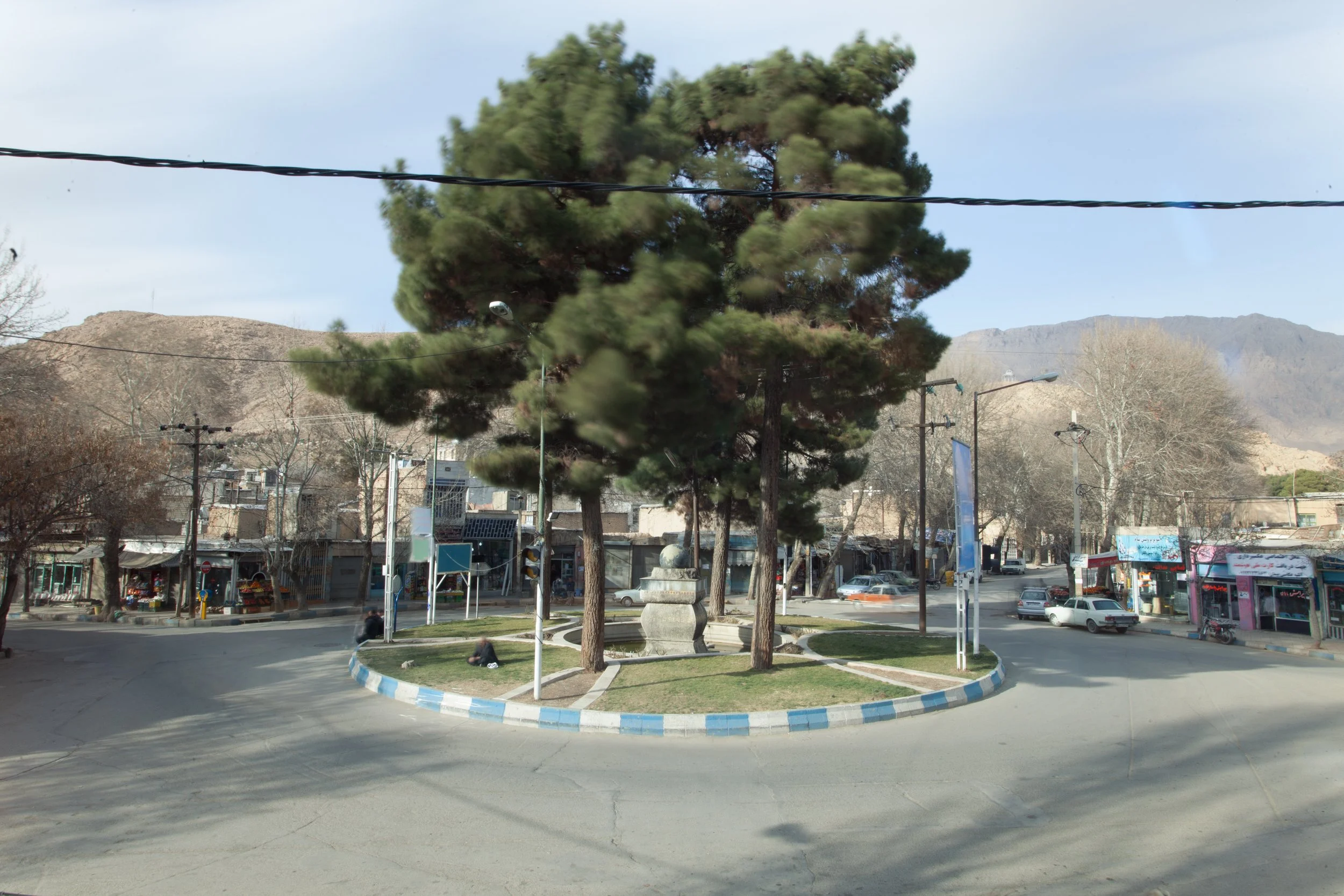 A small roundabout with a central monument surrounded by trees and grass, with cars and shops in the background, mountain hills behind.