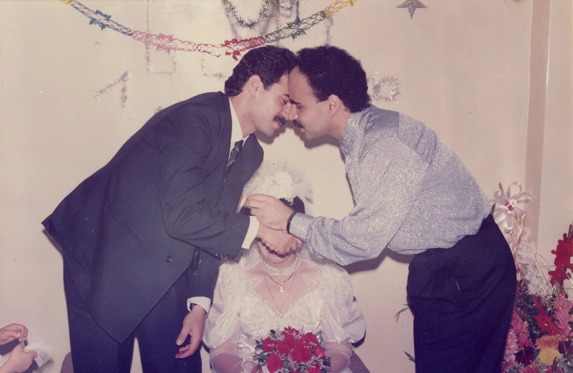 Two men in suits sharing a moment with an older woman in a wedding dress, likely at her wedding, with decorations and flowers in the background.