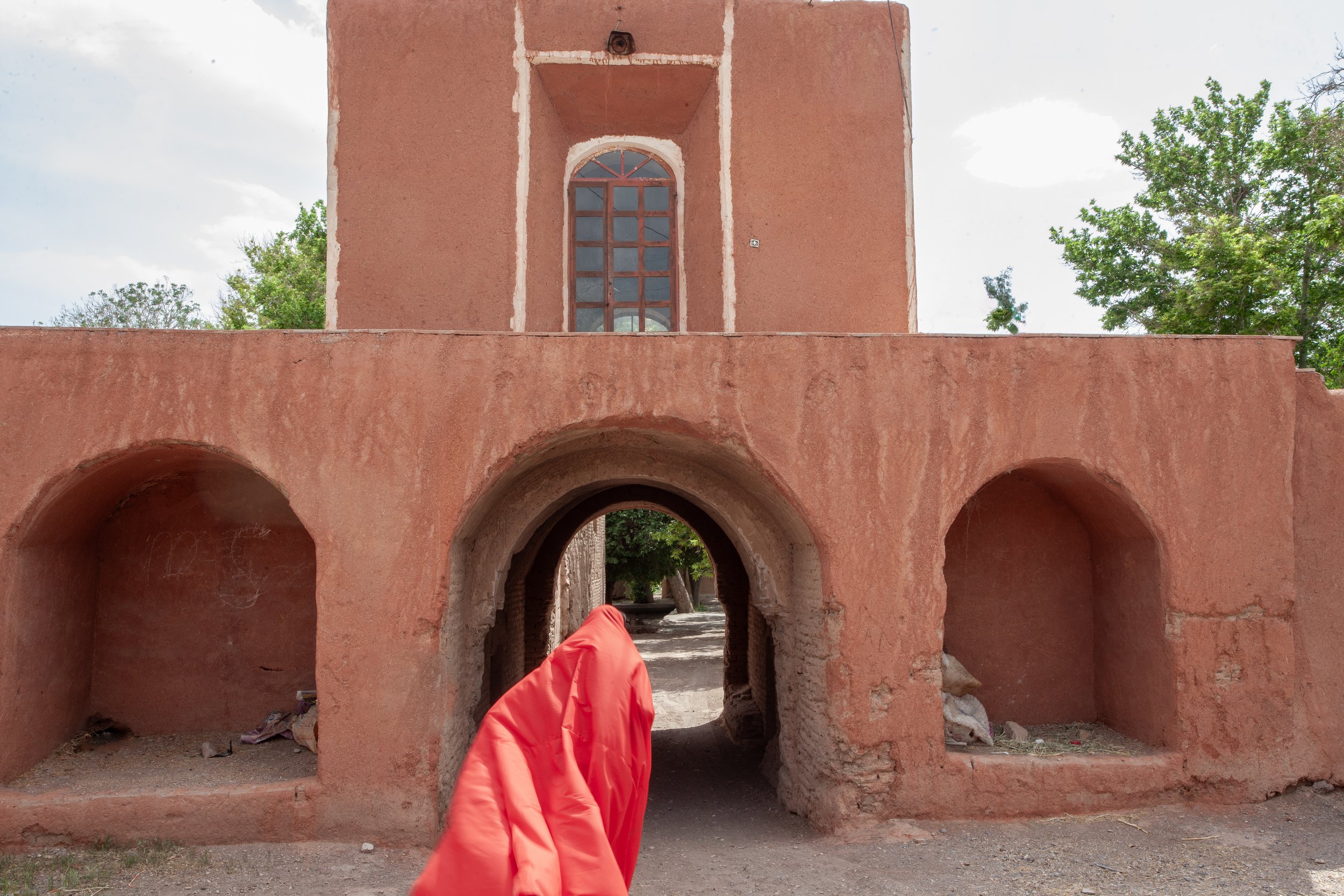 A pinkish adobe building with three arches in the front, and an arched window above the middle arch. There is a person dressed in a red windbreaker walking through the middle arch. Green trees are visible in the background.