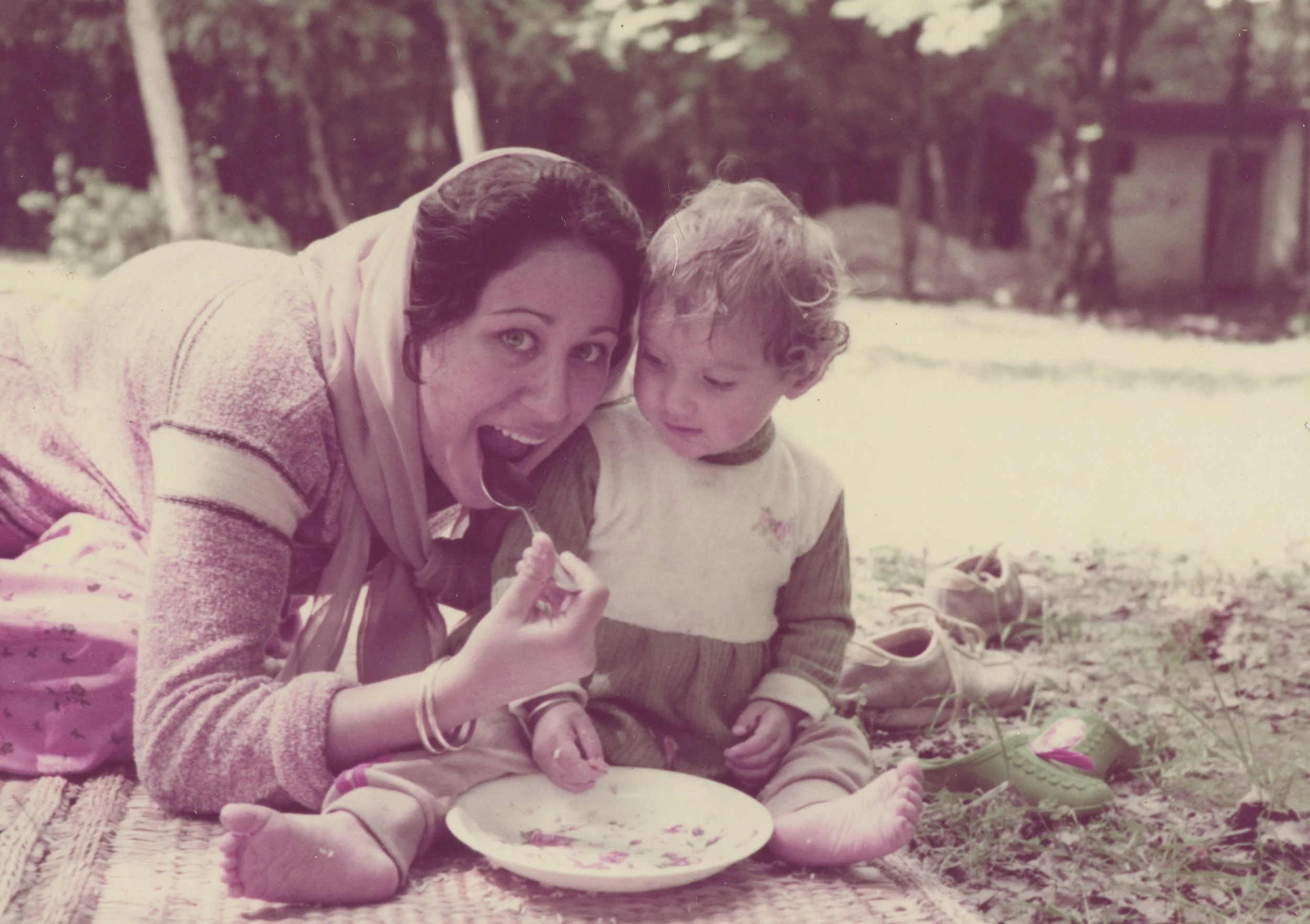 A woman and a young child sitting on a blanket outdoors, sharing a meal. The woman is holding a spoon, and there are shoes and a plate with food around them.