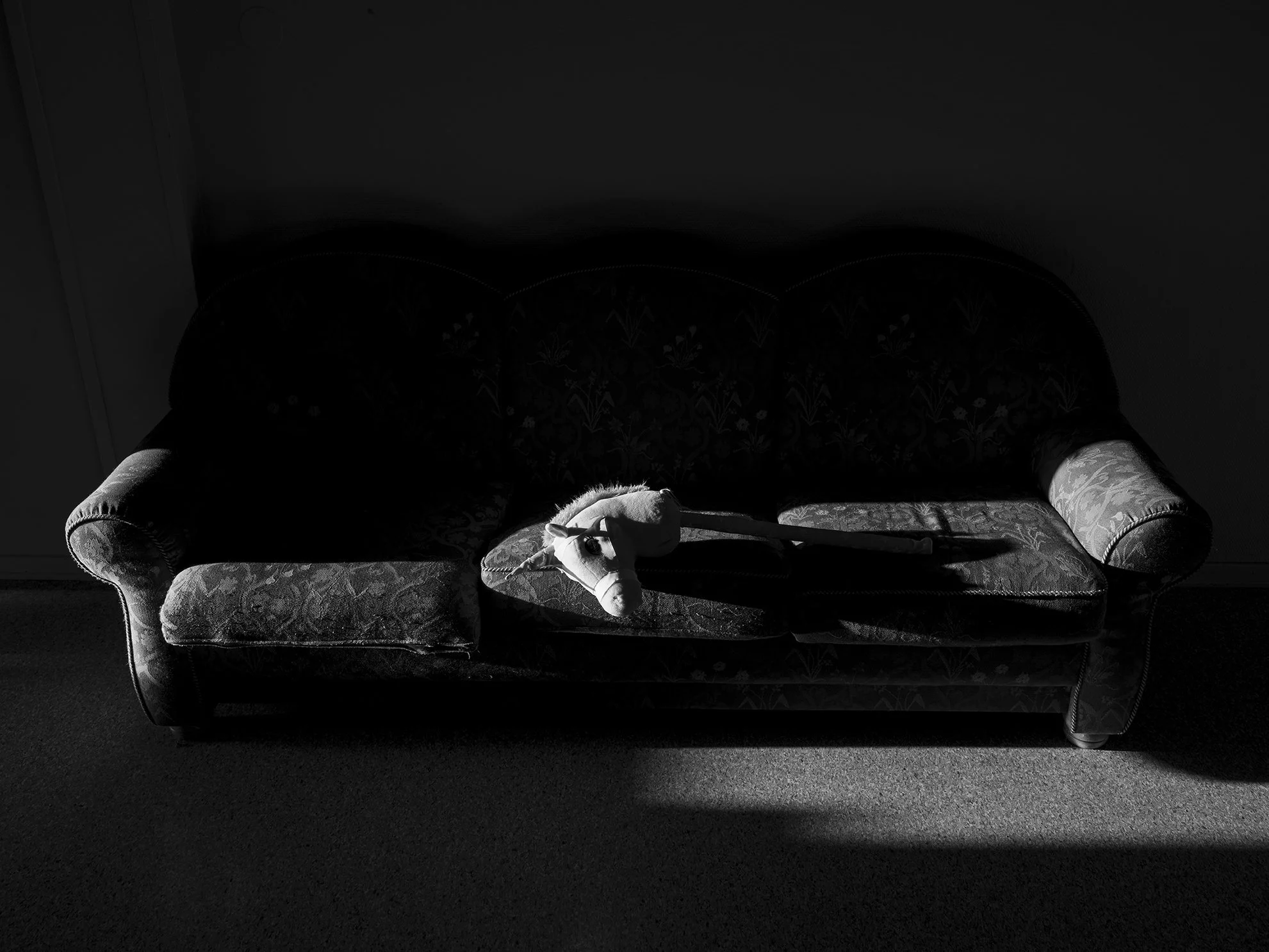 A vintage floral-patterned sofa with spilled upholstery and a plush horse toy lying on top, illuminated by a single beam of light in a dark room.