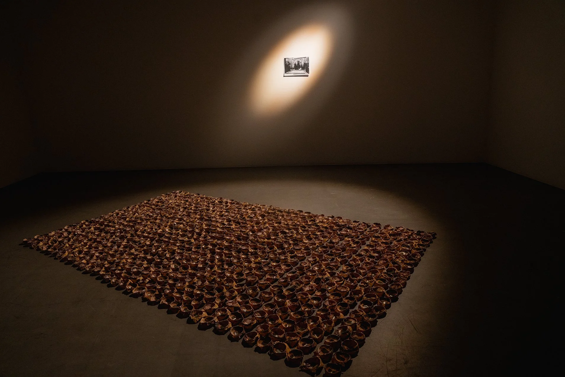 An art installation featuring a large square arrangement of cups or small containers on the floor in a gallery, with a spotlight illuminating a black-and-white photograph on the wall.