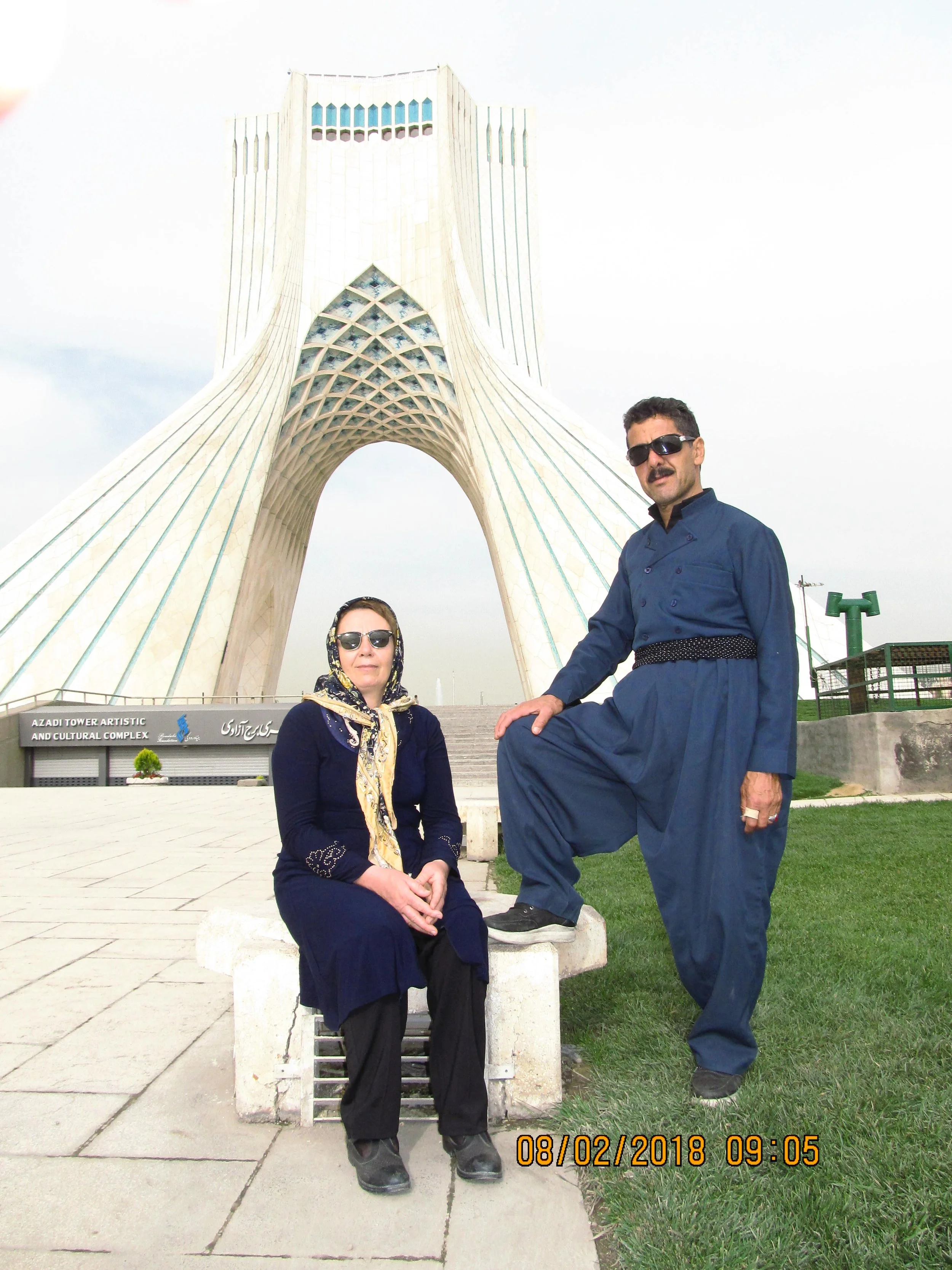A woman and a man posing in front of Azadi Tower, a tall, unique architectural monument. The woman is seated on a stone bench, wearing dark clothing, sunglasses, and a headscarf. The man is standing with one foot on a stone bench, wearing dark sunglasses and traditional Iranian attire. The scene appears to be at a cultural or tourist site.