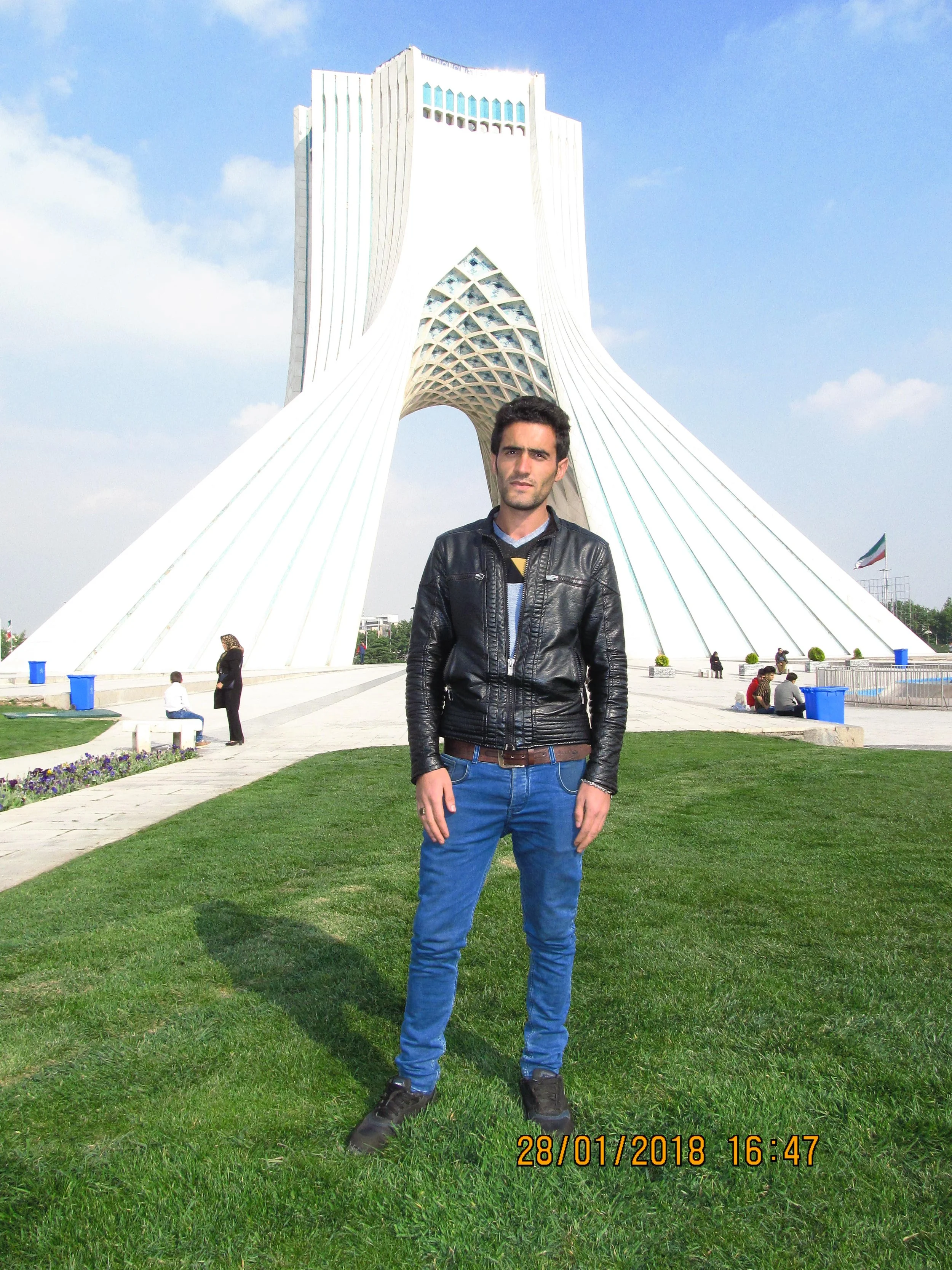 A man in a black leather jacket and blue jeans standing on green grass in front of Azadi Tower in Tehran, Iran, with a clear blue sky overhead.