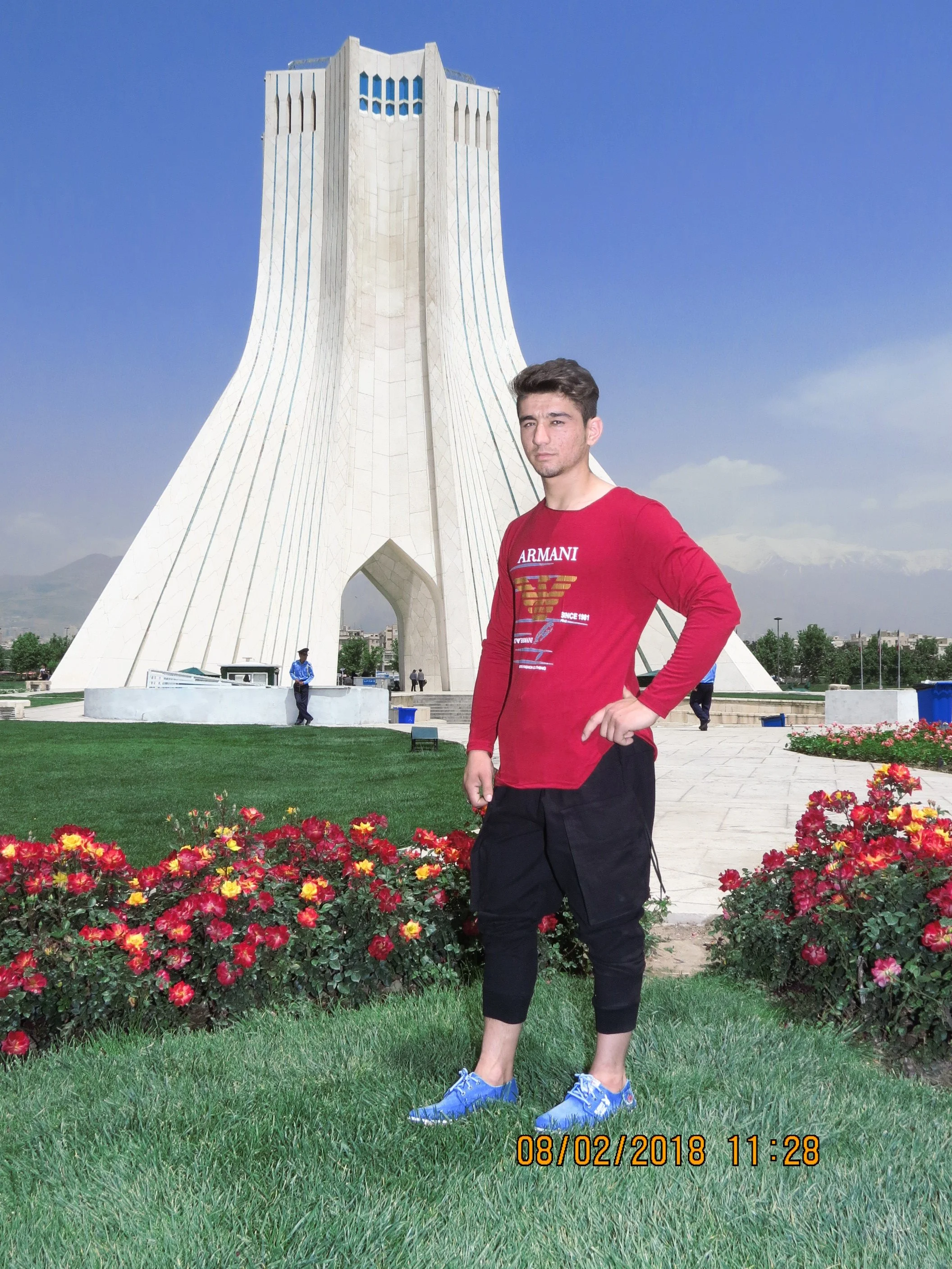 A young man posing outdoors in front of a large white monument, with green grass and pink flowers in the foreground, and distant mountains and blue sky in the background.