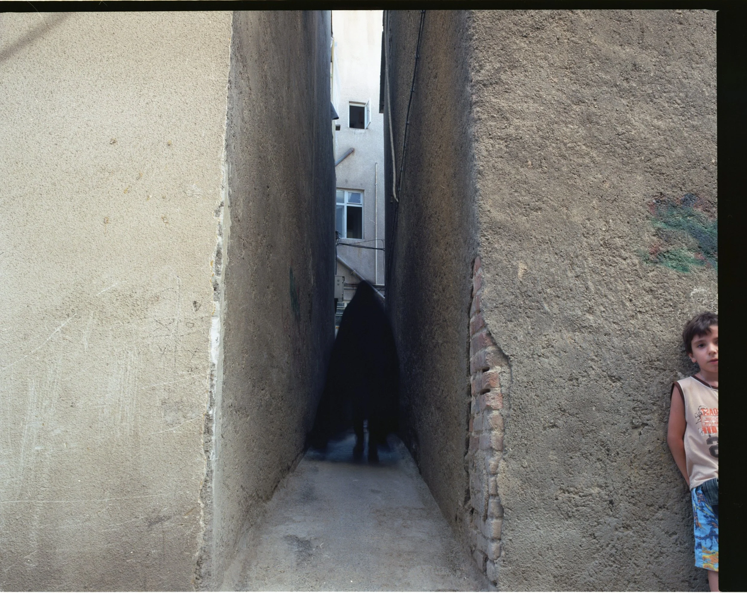 A narrow alleyway between two buildings with textured walls. There is a black dog in the shadows at the end of the alley, and a young boy with brown hair wearing a sleeveless shirt and colorful shorts leaning against the right wall.